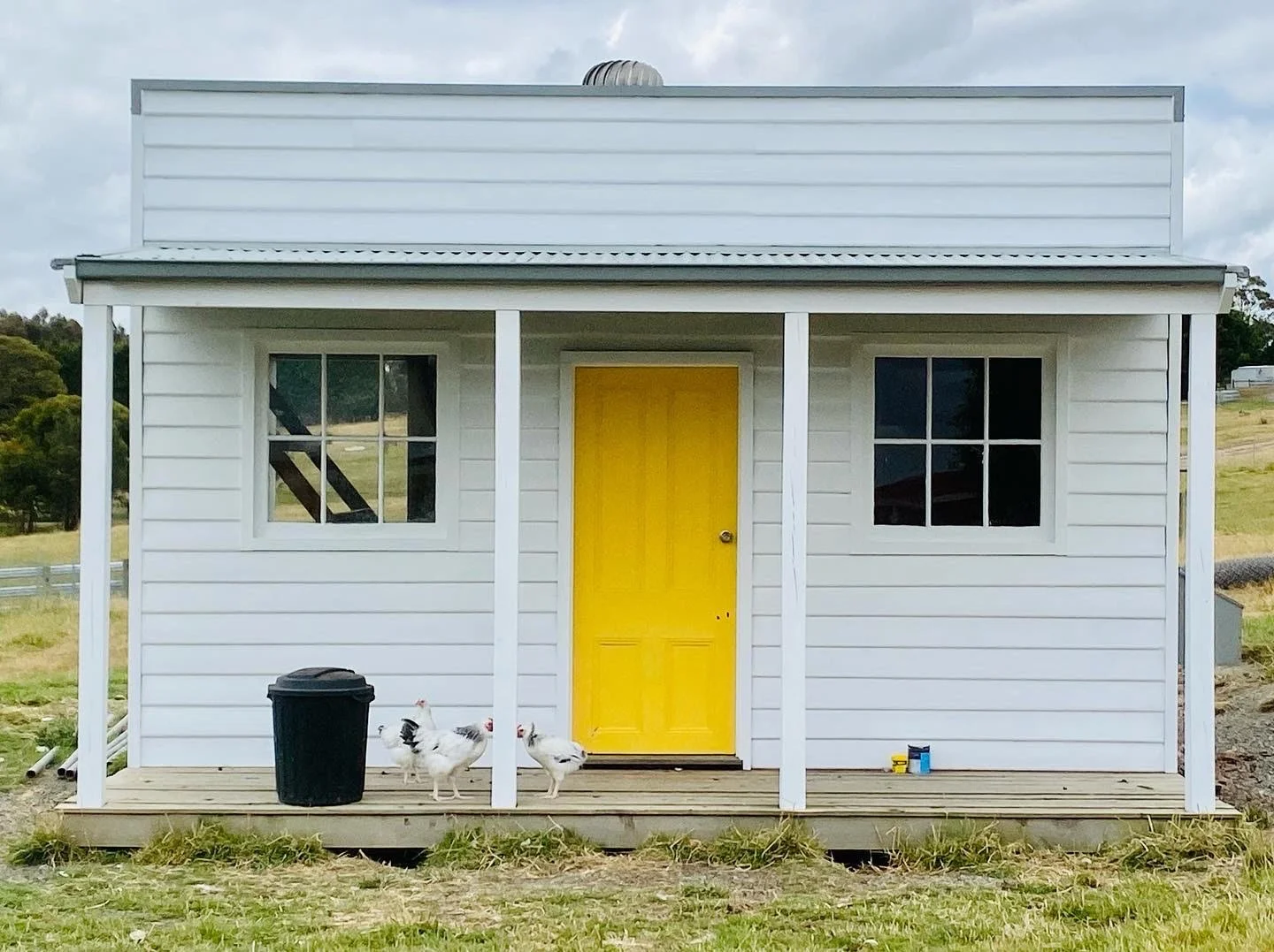 Small white house with a yellow front door, two windows, and a porch. Three chickens are on the porch, with a black trash can nearby. The house is on a grassy area with trees and hills in the background.