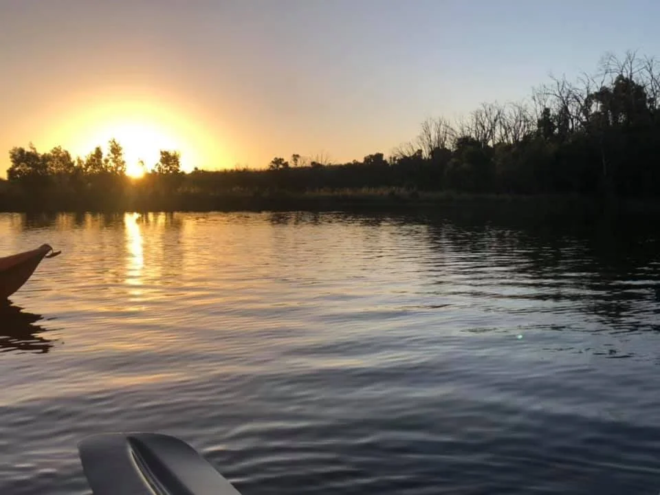 Sunset over a calm river with trees along the shoreline and a boat or canoe in the foreground.