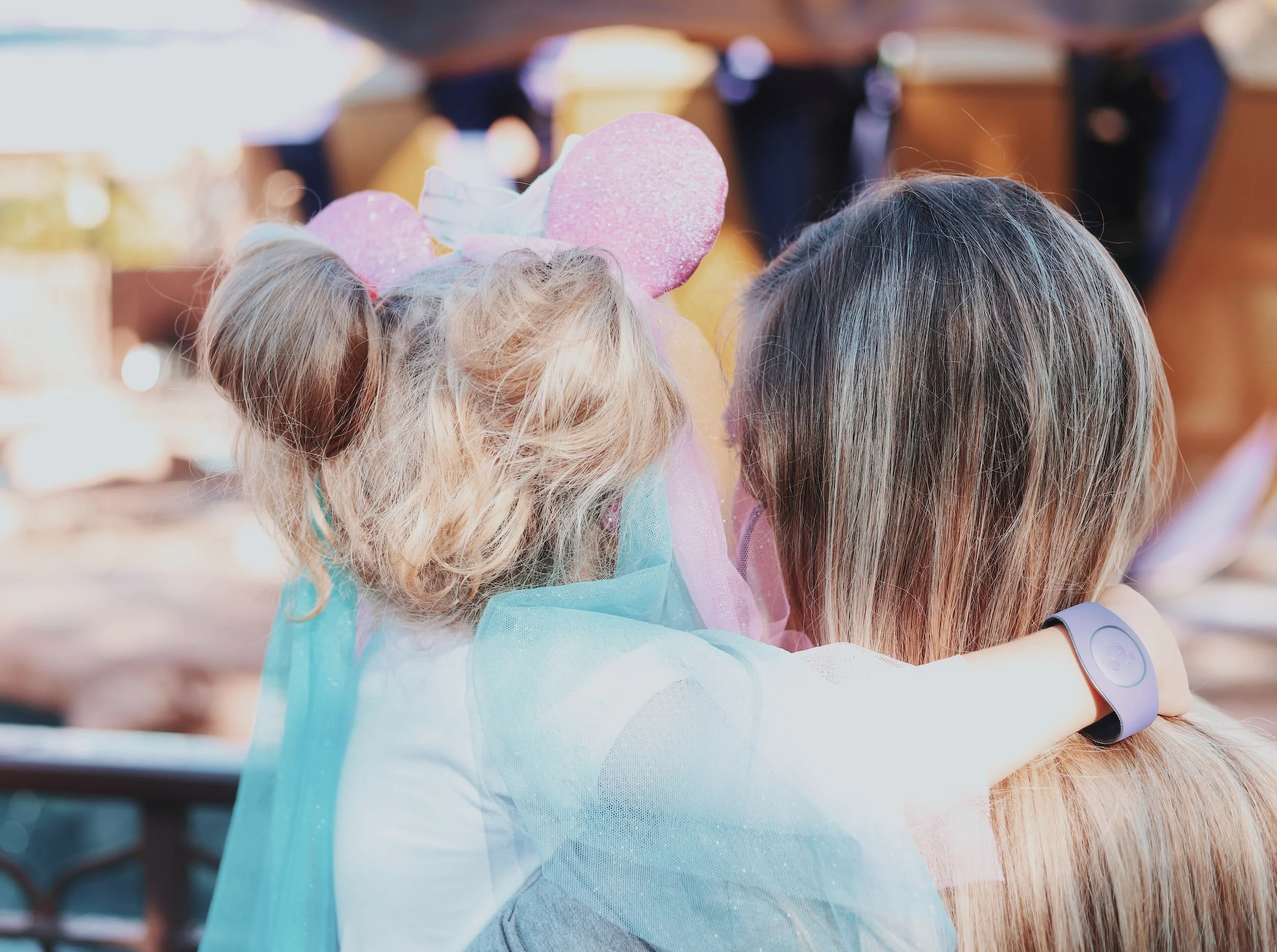 A young girl with Minnie Mouse ears and colorful tulle is hugging a woman with long hair, both seen from behind at an outdoor event.
