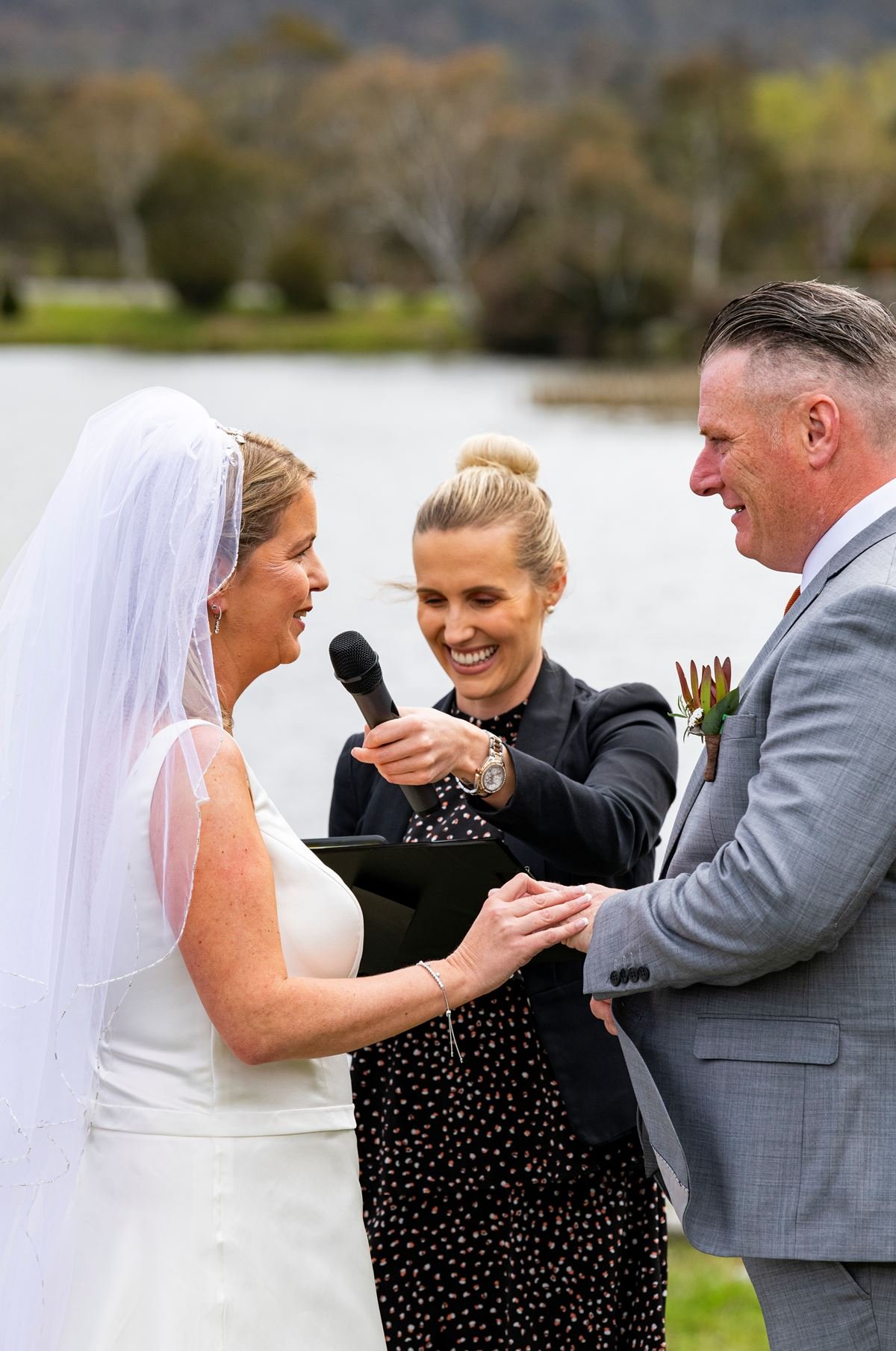 A bride and groom holding hands during an outdoor wedding ceremony, with a woman officiant speaking into a microphone near a body of water.