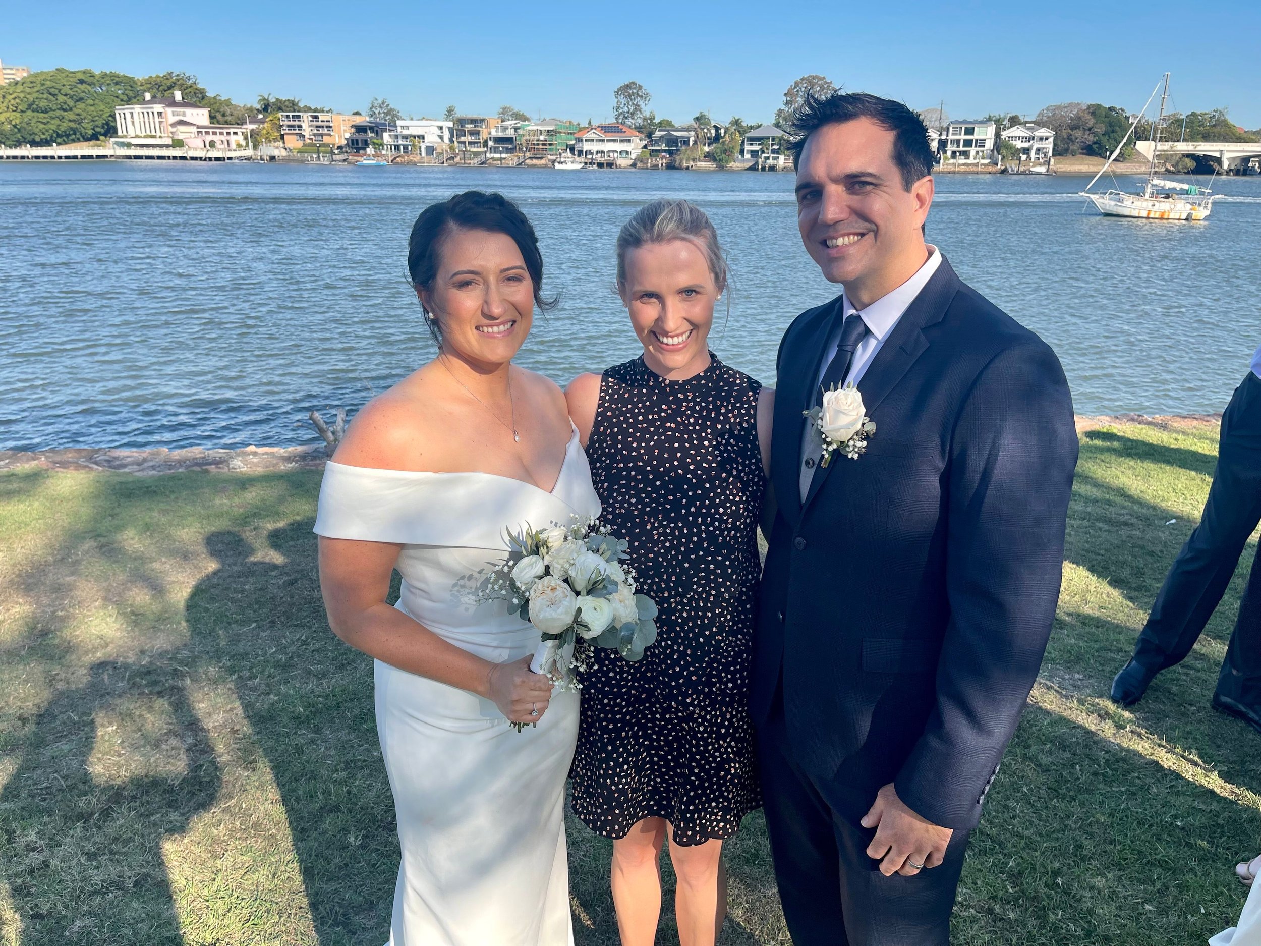 A bride and groom standing outdoors by a body of water with a woman in a black dress between them, all smiling at the camera.