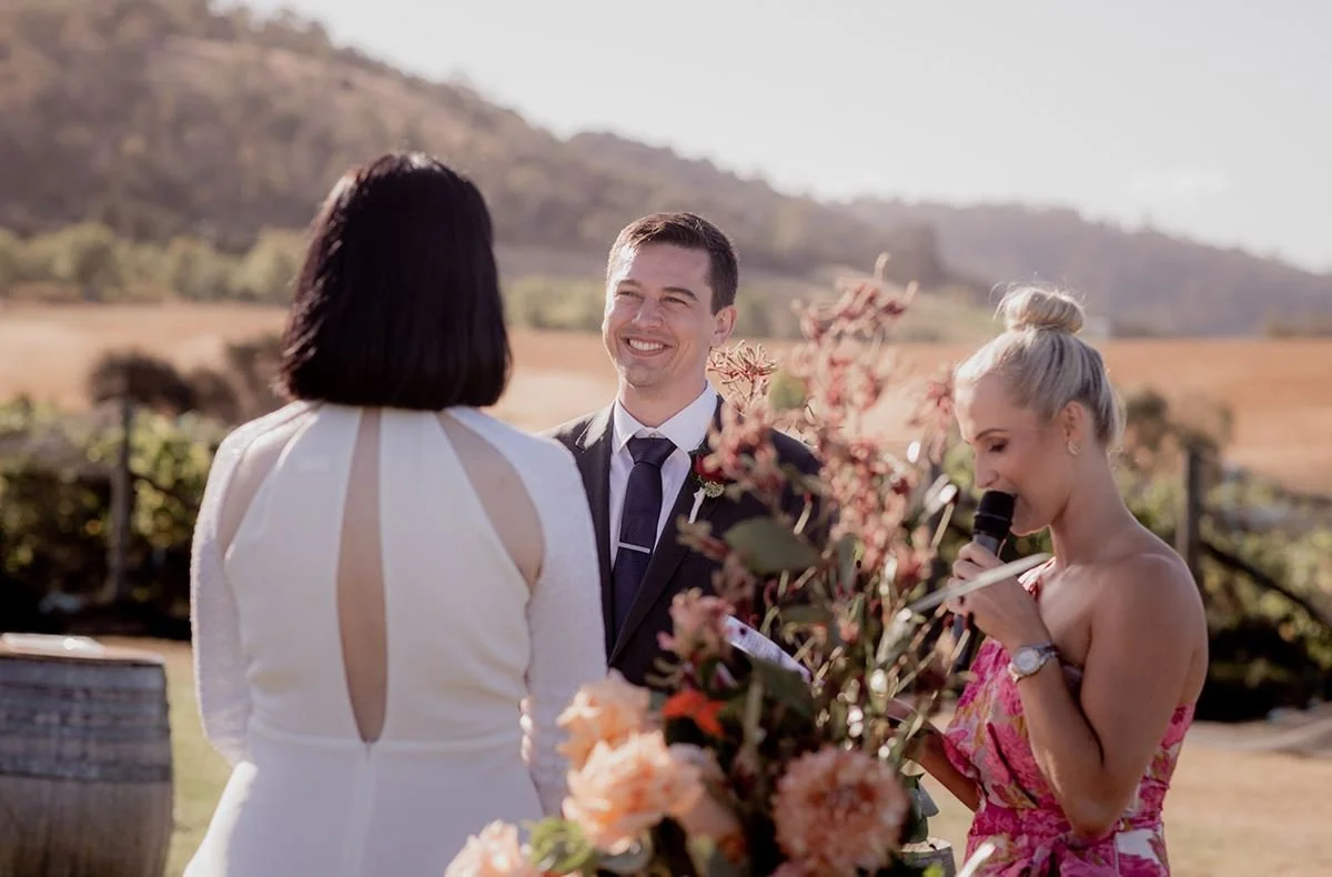 A wedding ceremony outdoors with a officiant reading from a paper and the groom smiling, surrounded by flowers, with hills in the background.