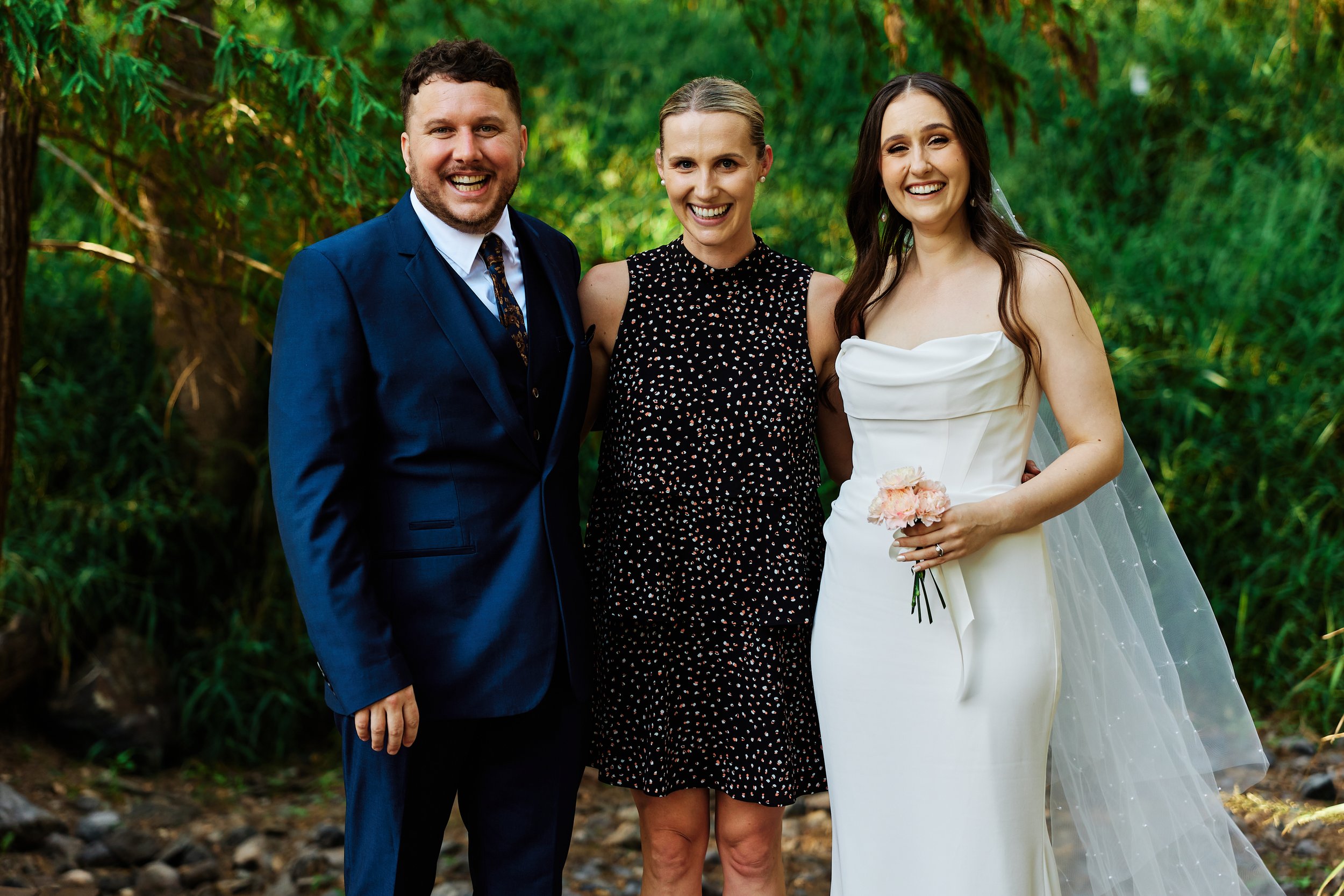 A wedding celebration outdoors featuring a man in a blue suit, a woman in a black polka dot dress, and a bride in a white wedding gown holding pink flowers, all smiling.