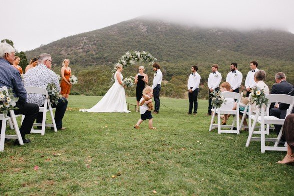 Outdoor wedding ceremony with bride, groom, officiant, and guests on a grassy field with mountains in the background