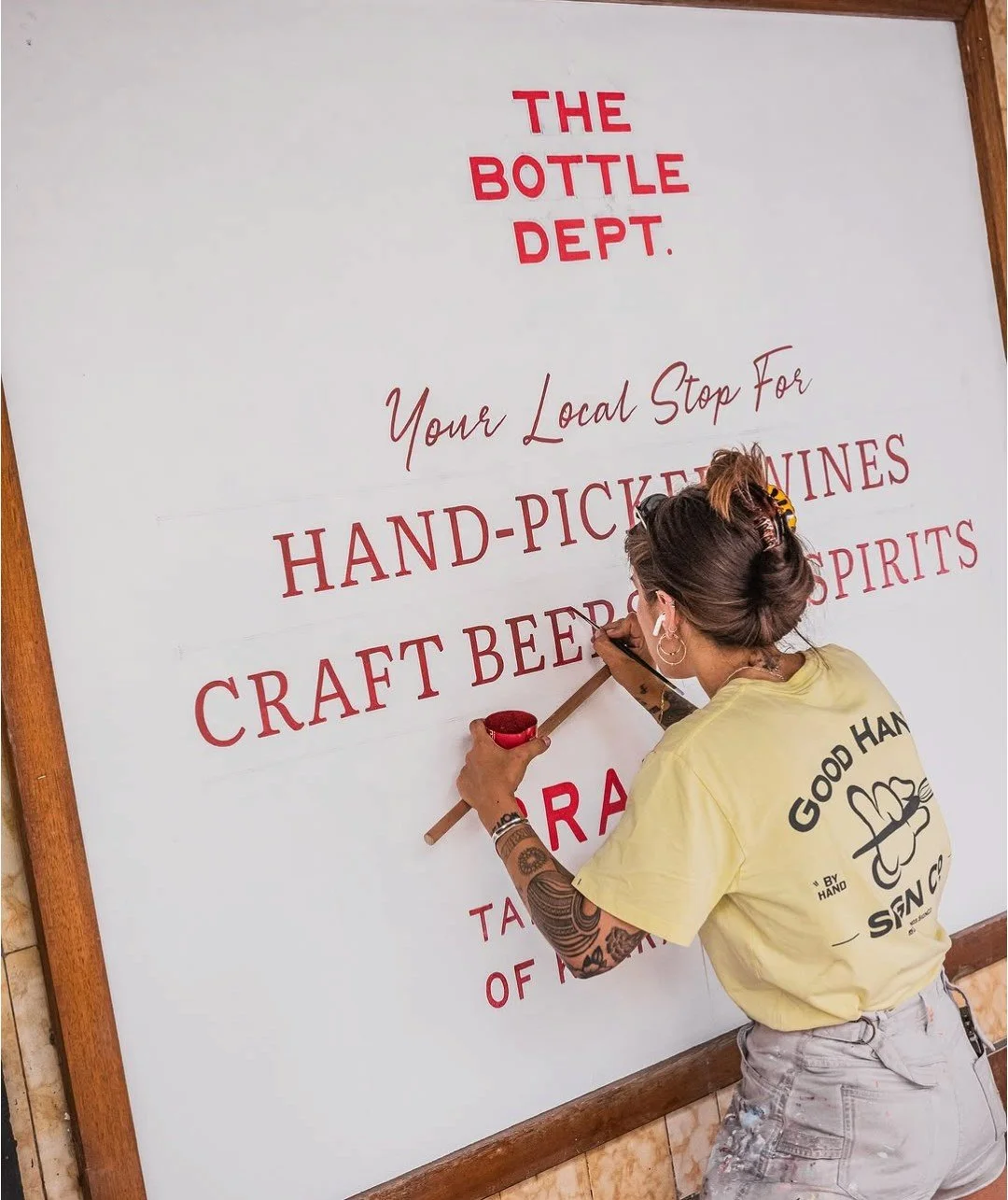 A woman is painting a sign on a large whiteboard with red and brown text, wearing a yellow T-shirt and gray pants, with tattoos on her right arm, in a wooden-framed room.