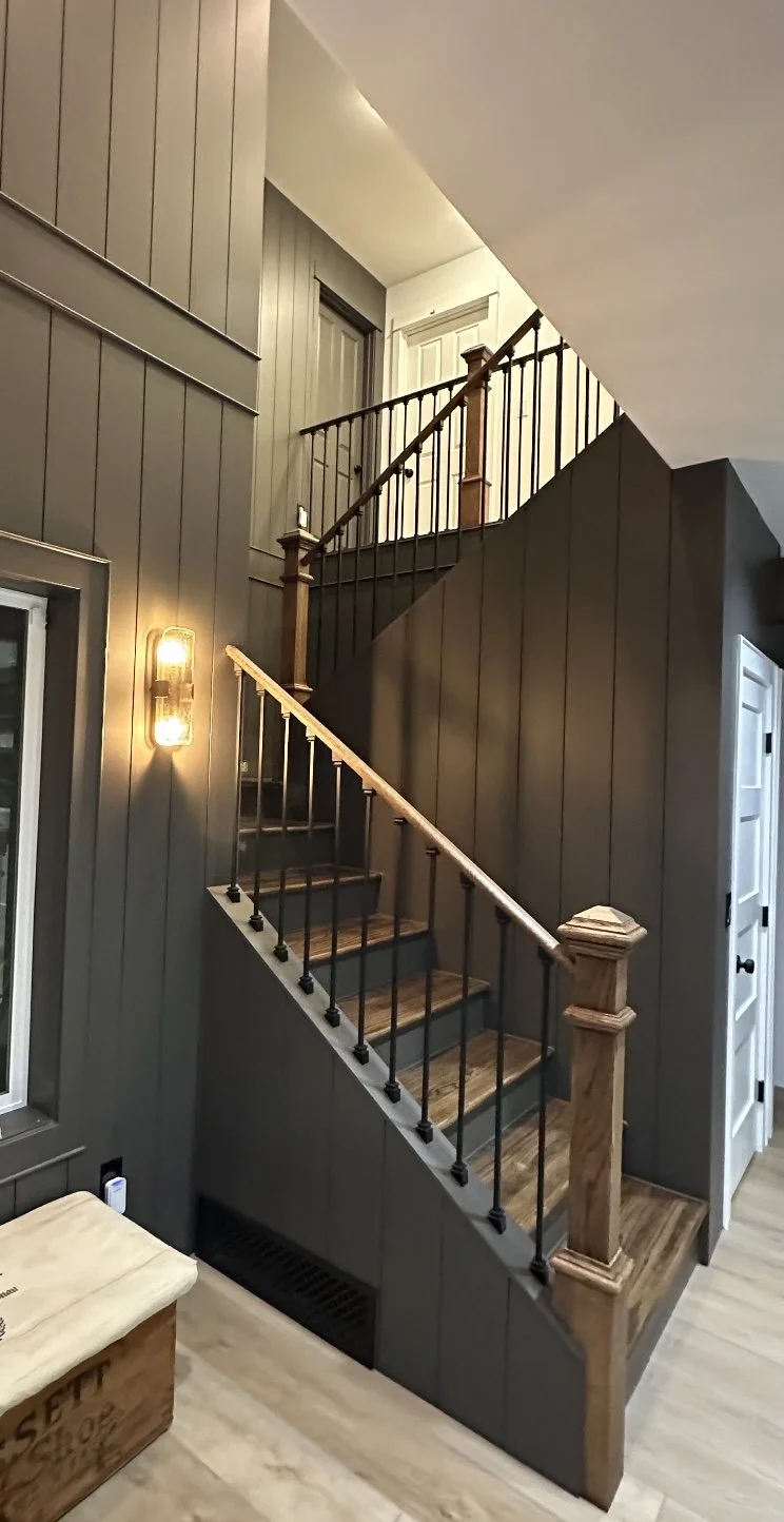 Interior of a home showing a staircase with wooden steps and black metal balusters, dark gray paneled walls, a wall-mounted light fixture, and a white door at the top of the staircase.