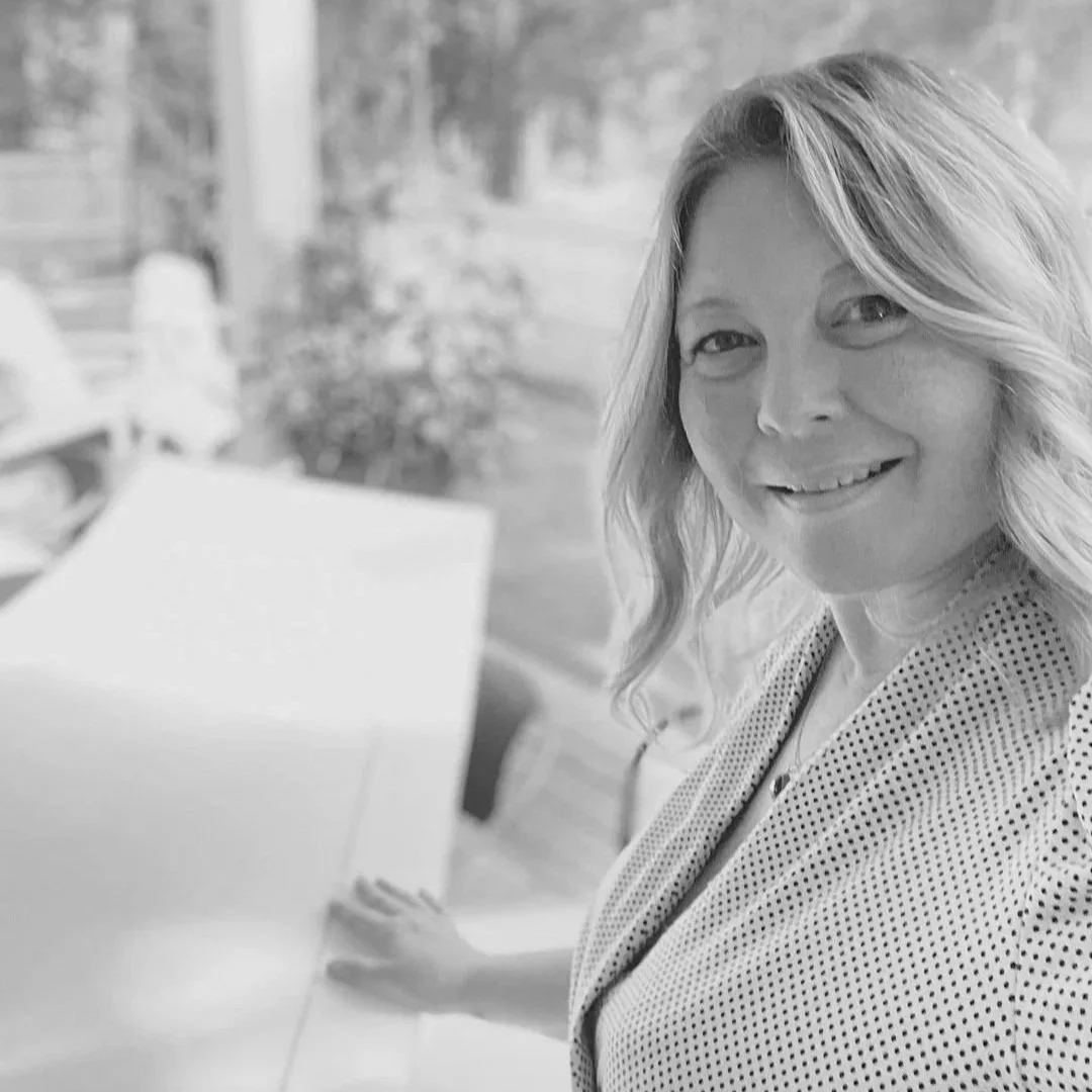 A woman with wavy hair smiling while sitting at a table outdoors in black and white.