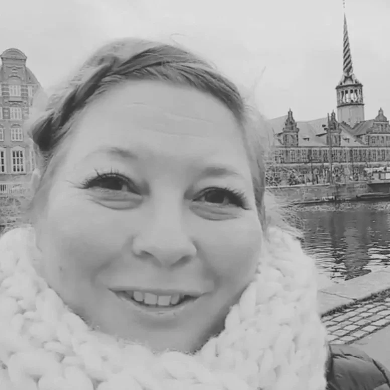 A woman smiling near a body of water, with historical European-style buildings and a tall spire in the background.