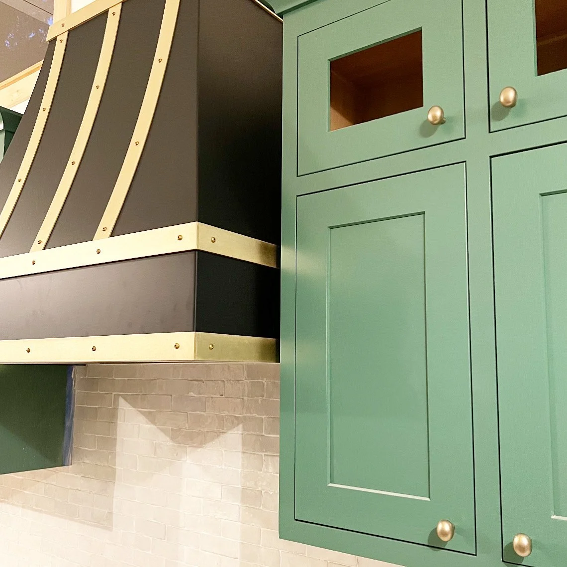 Close-up of green kitchen cabinet with two doors and round silver knobs, beside a black and beige structure with gold trim and rivets, and a beige brick wall.
