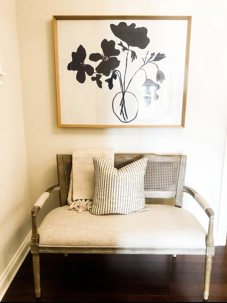 A beige upholstered bench with wooden armrests and backrest, topped with a striped pillow and a textured beige blanket, in front of a white wall with a large framed black and white botanical print of flowers in a vase.