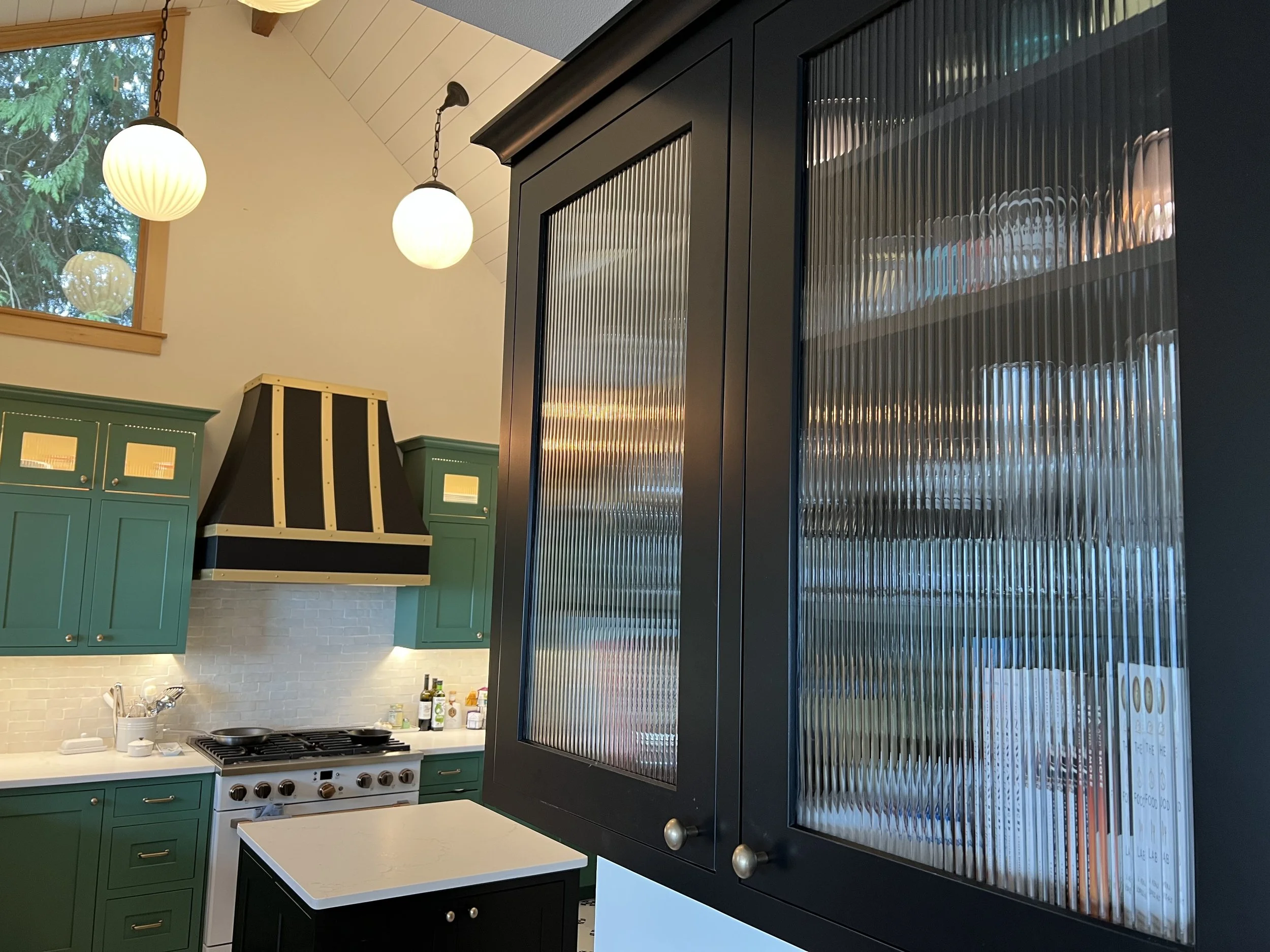 Modern kitchen with green cabinets, a black range hood, white countertops, and a large black cabinet with textured glass doors. Two spherical pendant lights hang from the ceiling near a window.