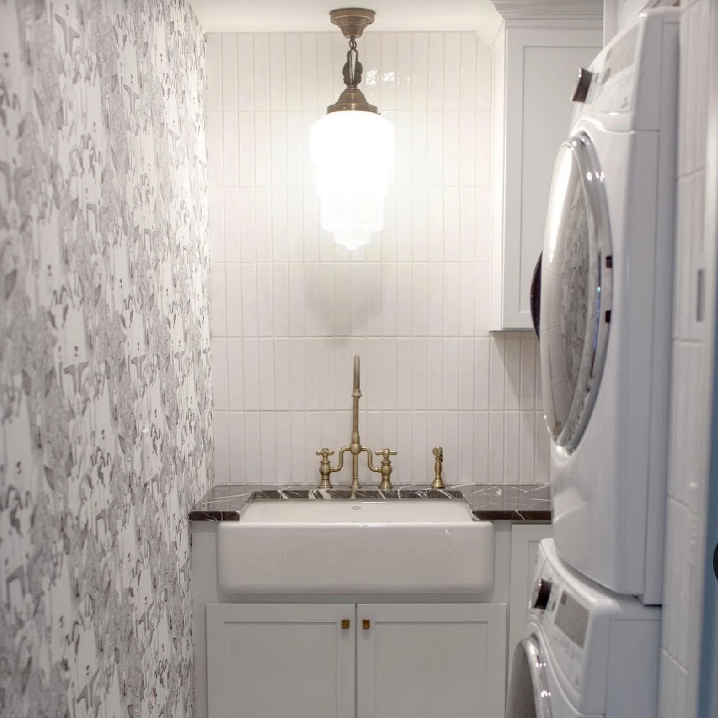 Laundry room with stacked washer and dryer on the right, a utility sink with brass faucet in the center, white cabinets above and below, a hanging light fixture, and a wall with patterned wallpaper on the left.
