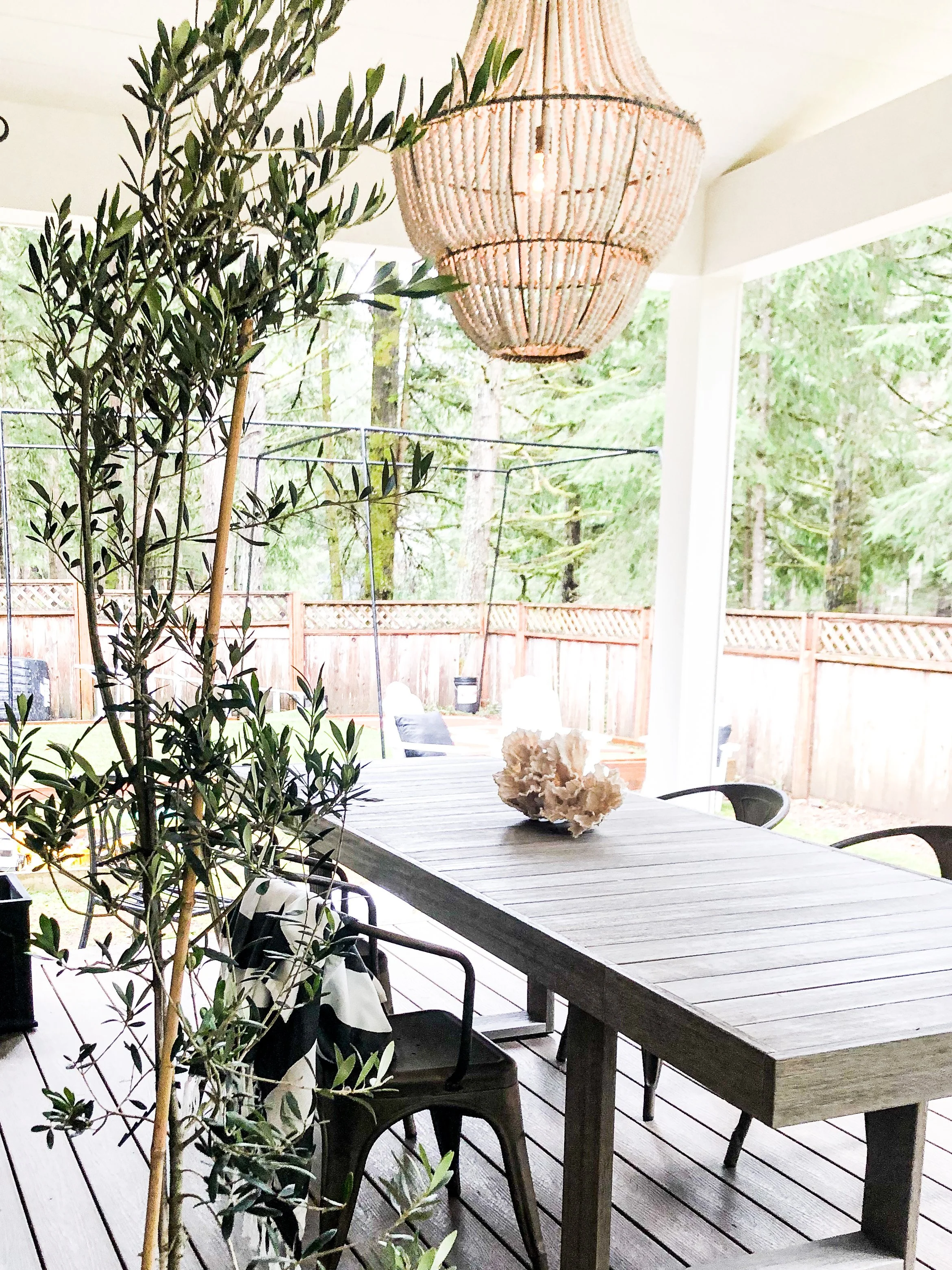 Outdoor patio with wooden dining table, black chairs, greenery, and a large woven chandelier.