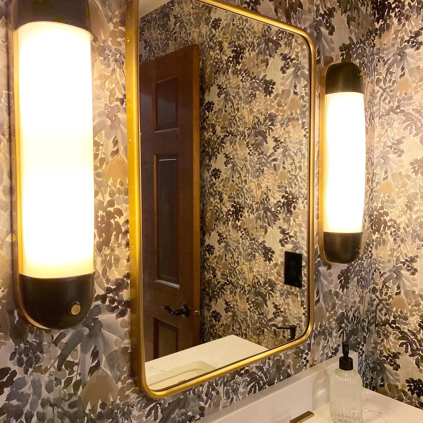 Bathroom with floral wallpaper, a large gold-framed mirror, wall-mounted oval black and white lights, a wooden door, and a sink with a clear soap dispenser.