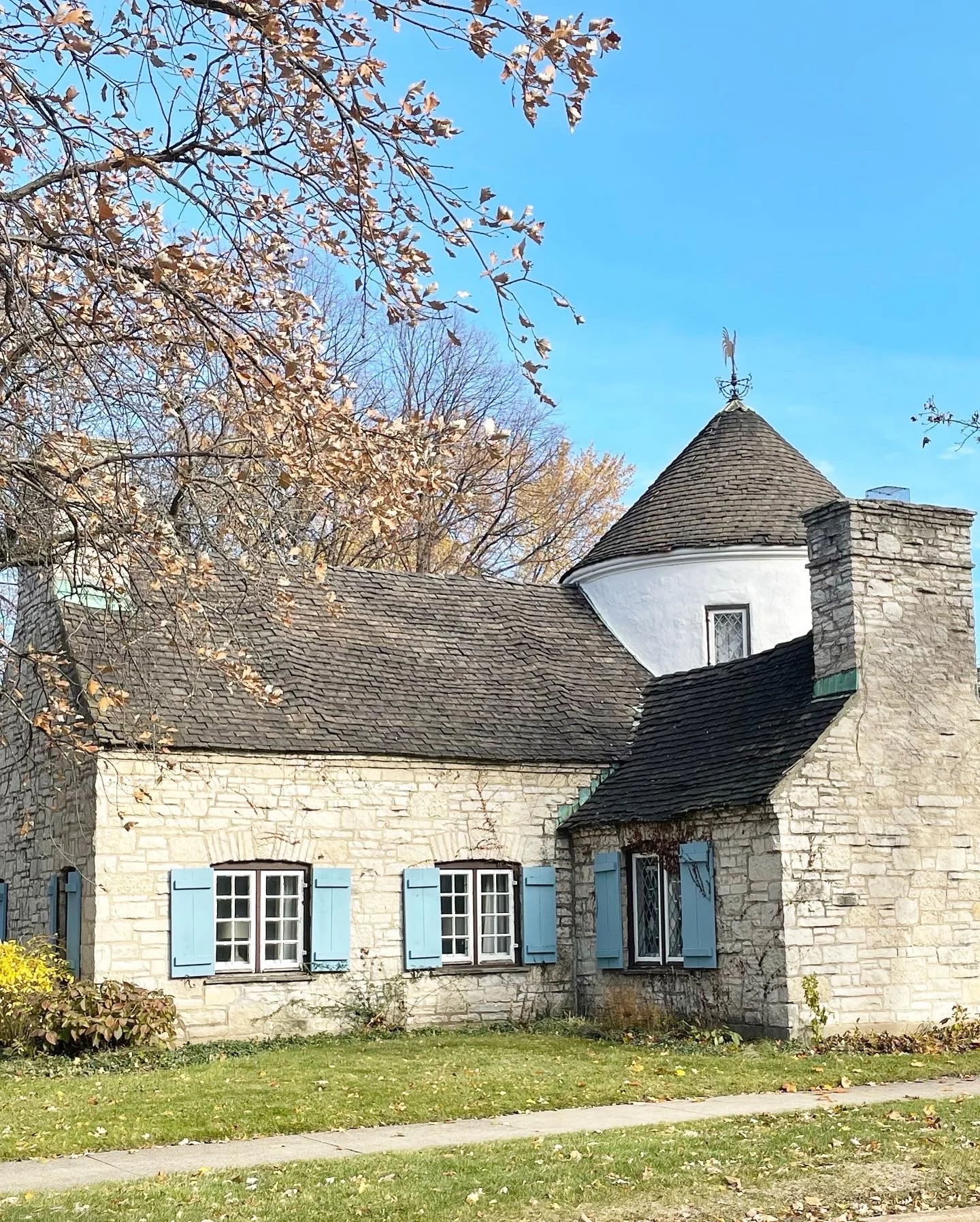 A quaint stone house with blue shutters on the windows, a grassy front yard, and a conical turret with a weather vane. There are trees with autumn leaves in the background and a clear blue sky.