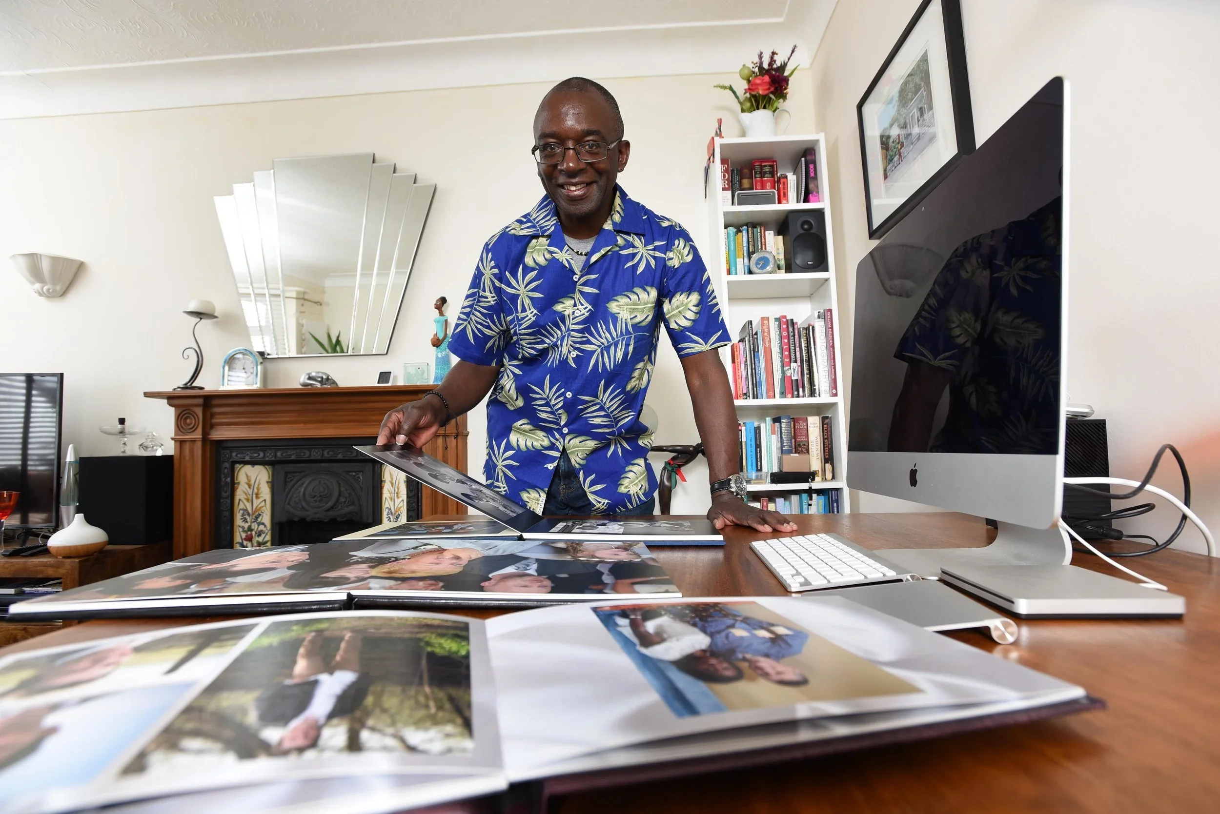 A man in a blue tropical shirt stands at a desk looking at photo albums. The desk has an iMac computer, a keyboard, and scattered photos. A bookshelf, mirror, and fireplace are in the background.