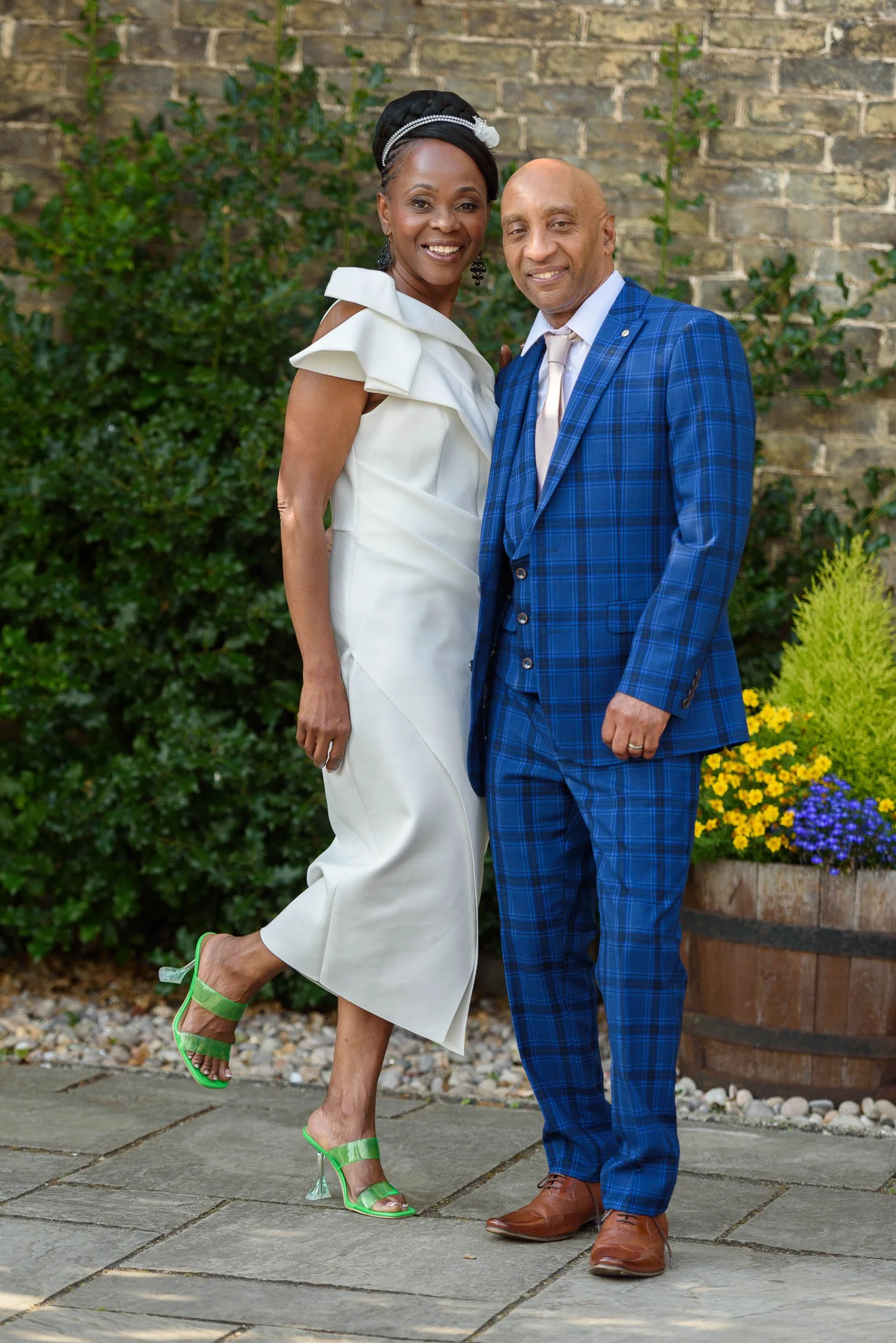 A woman in a white dress and green high heels, and a man in a blue checkered suit, standing outdoors in front of a brick wall and greenery, smiling at the camera.