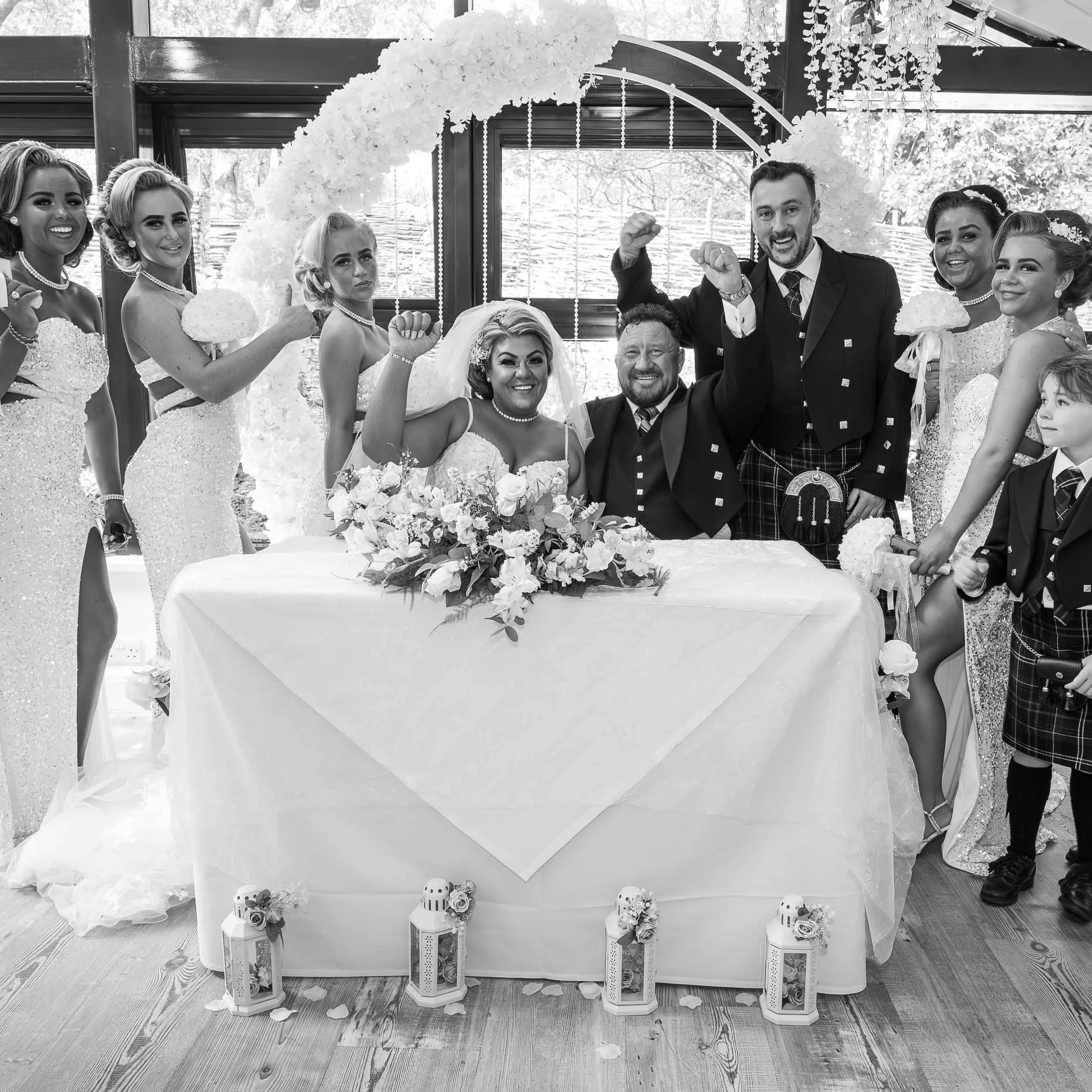A black and white photo of a wedding celebration at a table decorated with flowers and lanterns, featuring a bride and groom seated and celebrating with family and friends.
