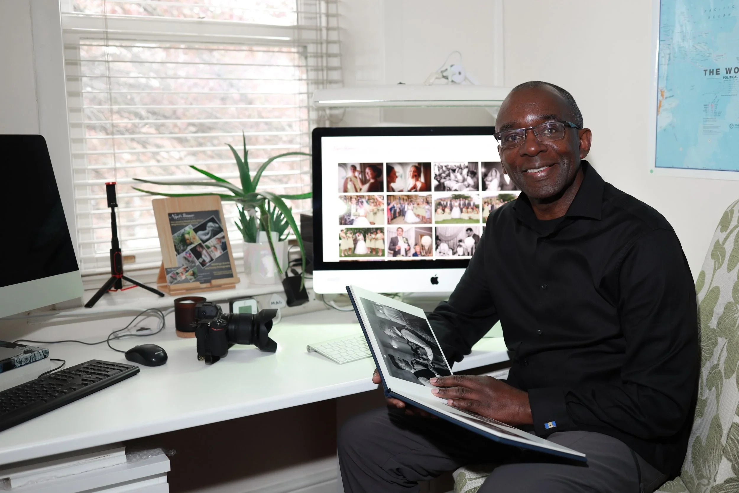 A man sitting in an office during daytime, holding a photo album and smiling at the camera. Behind him there is a computer monitor displaying photos, a potted plant, and various desk items including a camera and a keyboard.