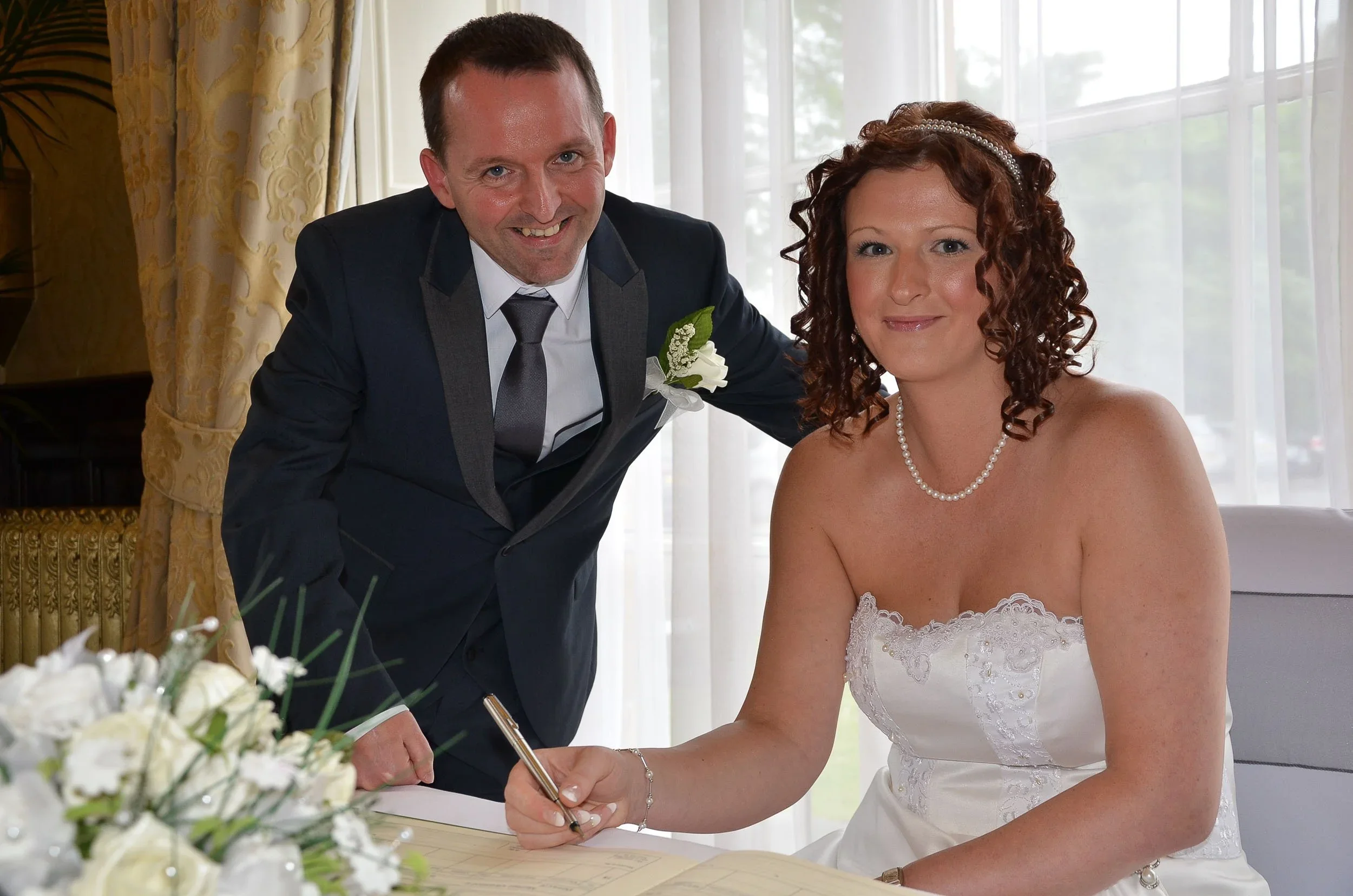 A bride with curly brunette hair wearing a pearl necklace and a strapless white wedding dress is sitting and signing a document, while a smiling man in a dark suit and gray tie, with a white flower boutonniere, leans over her in a well-lit room with large windows and yellow curtains.