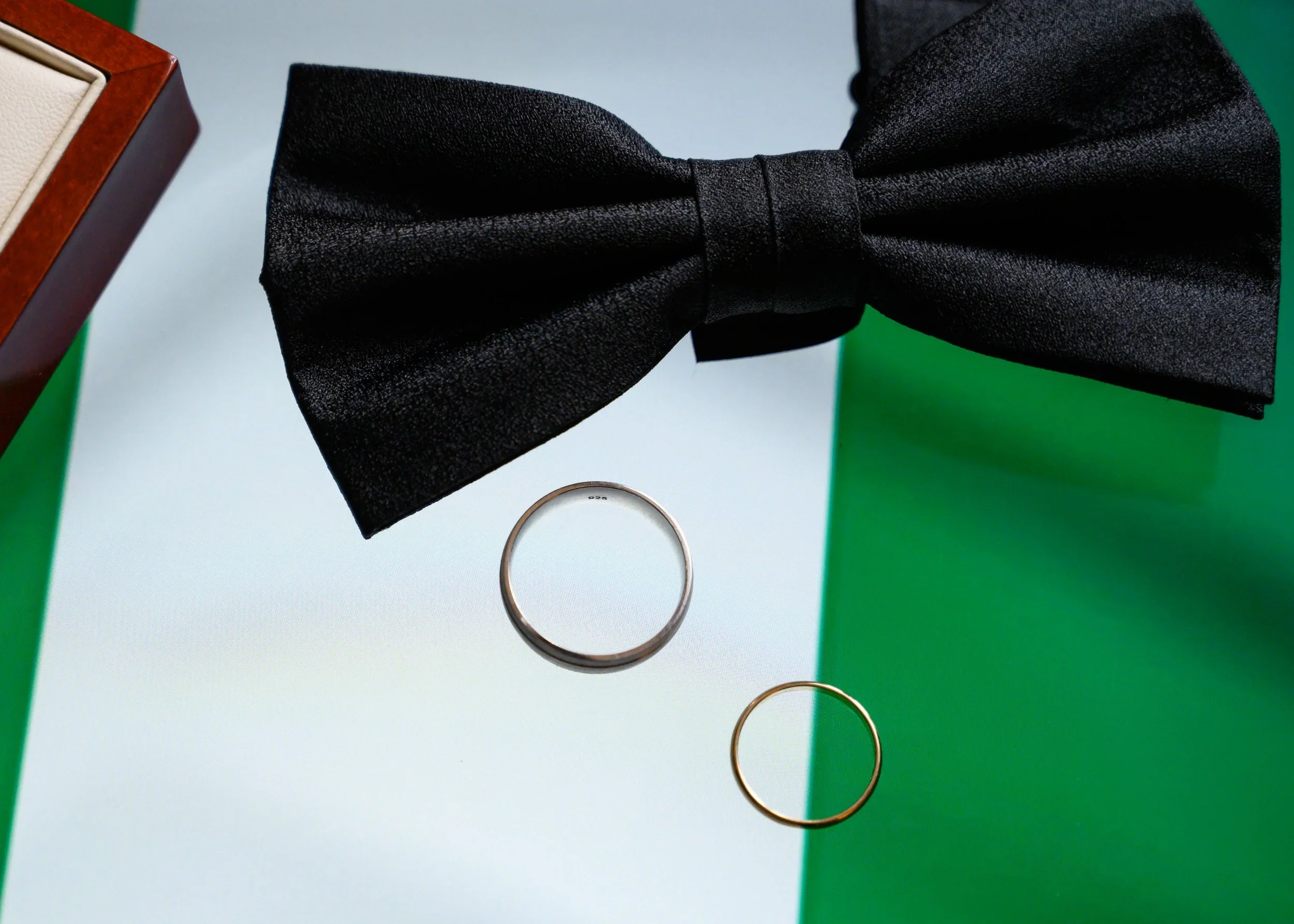 Close-up of a black bow tie, two rings (one silver, one gold), a jewelry box, and a green and white background.