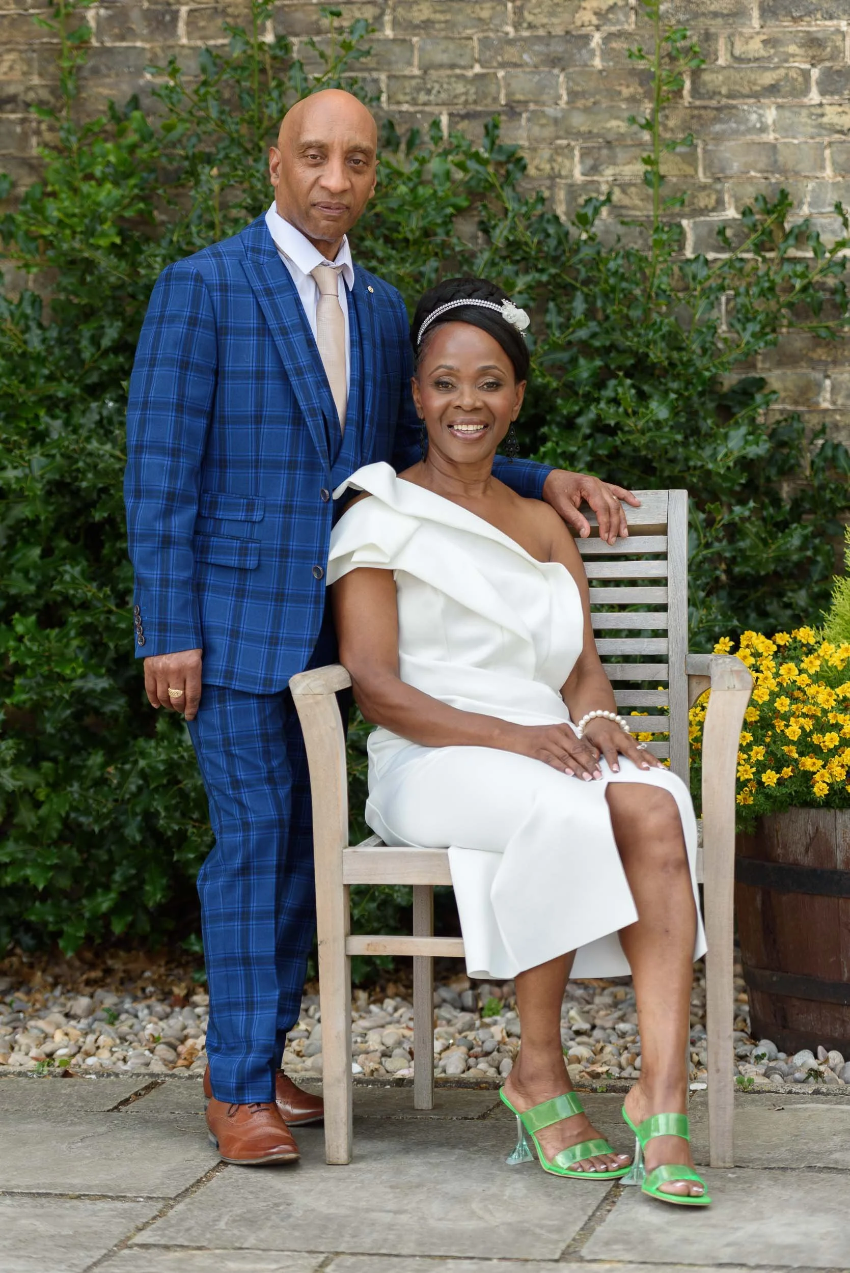 A man and a woman, dressed formally, pose outdoors with greenery and a brick wall in the background. The woman is seated on a wooden chair, wearing a white dress with a ruffled shoulder detail, green heels, and a pearl bracelet. The man is standing beside her, wearing a blue plaid suit with a light-colored tie and brown shoes.