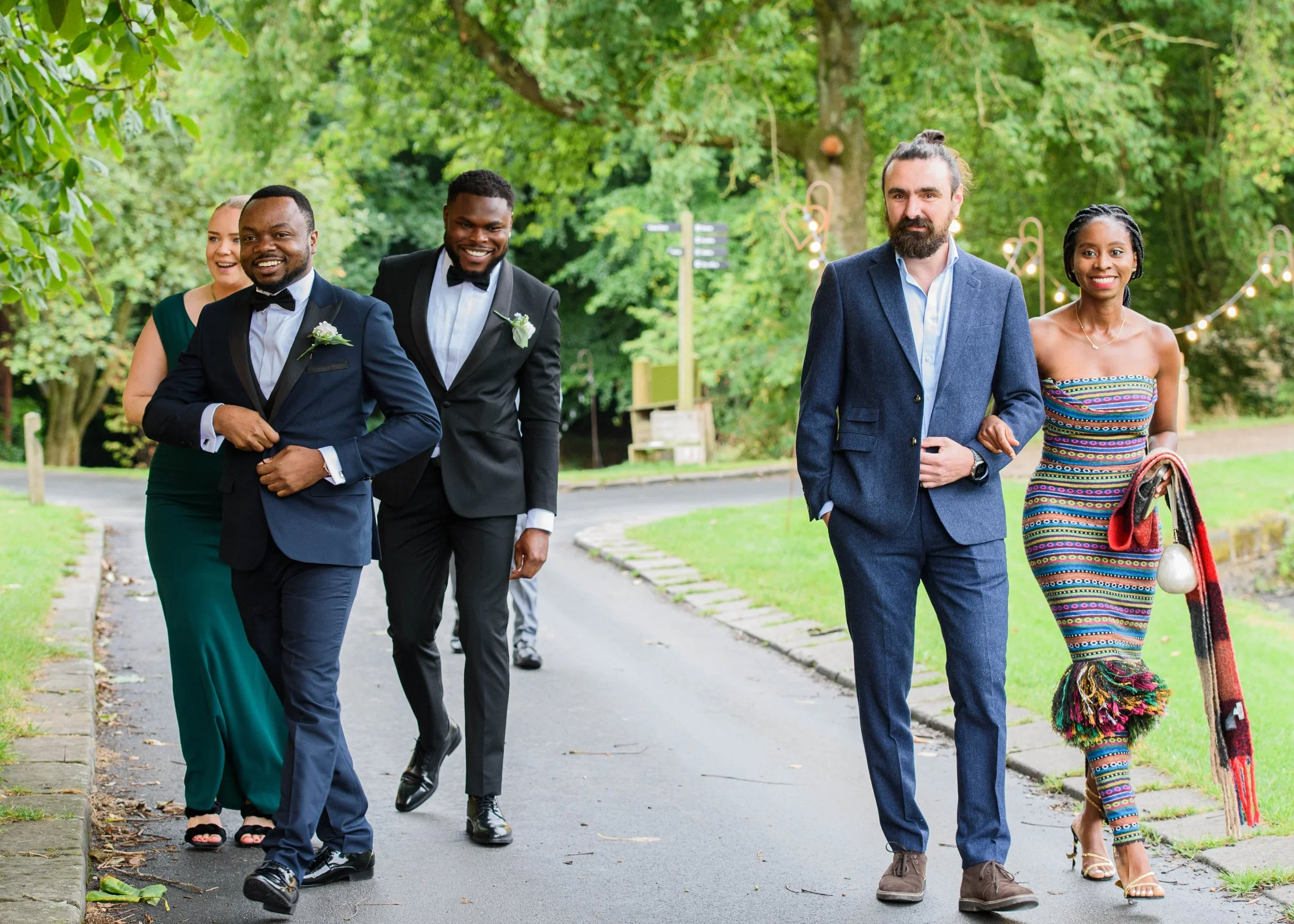 Group of five people walking outdoors on a paved path, dressed in formal and casual attire, with trees and string lights in the background.