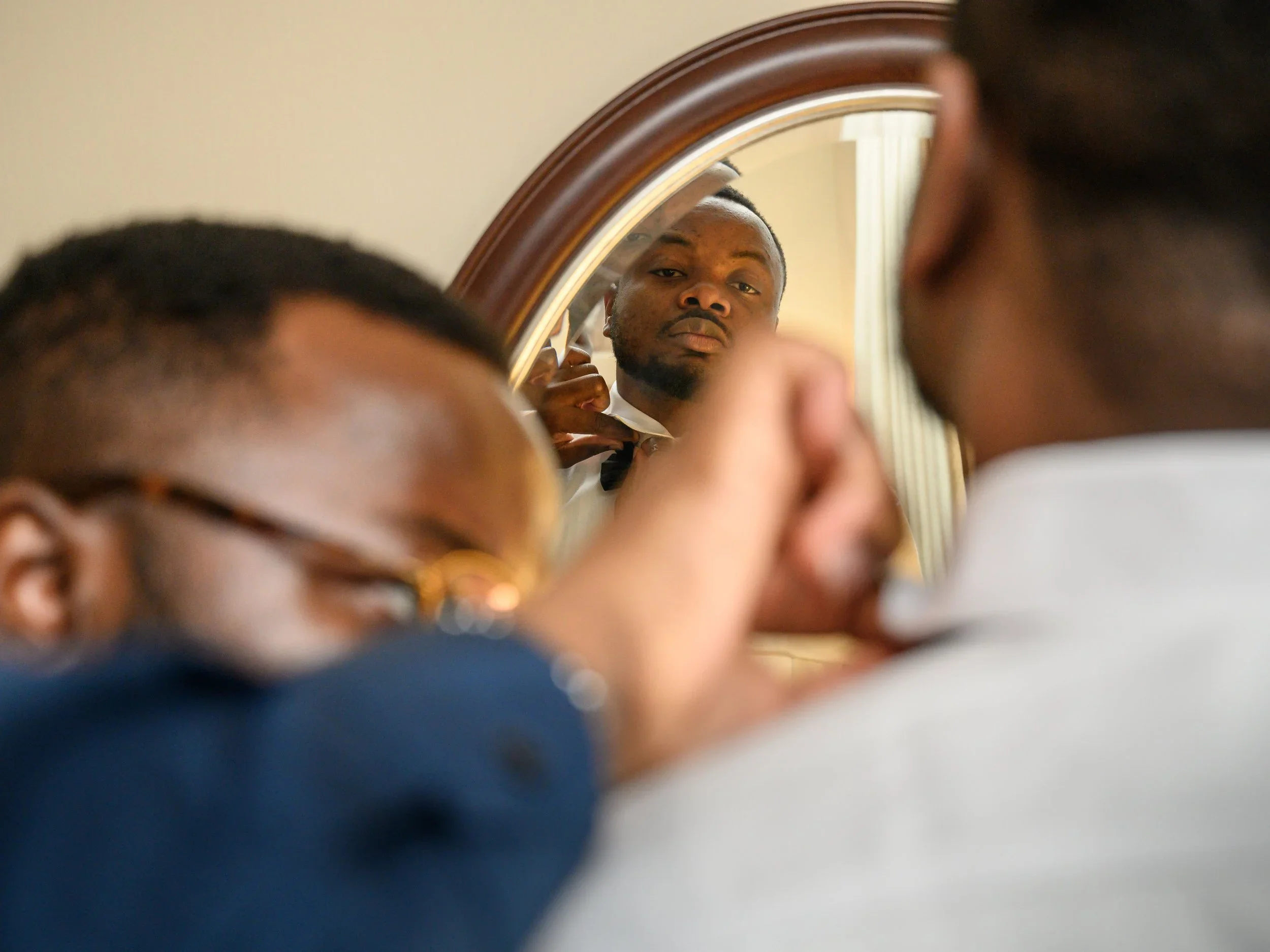 photo of a black groom standing in front of a mirror, having his bow tie fixed by another black man wearing glasses.
