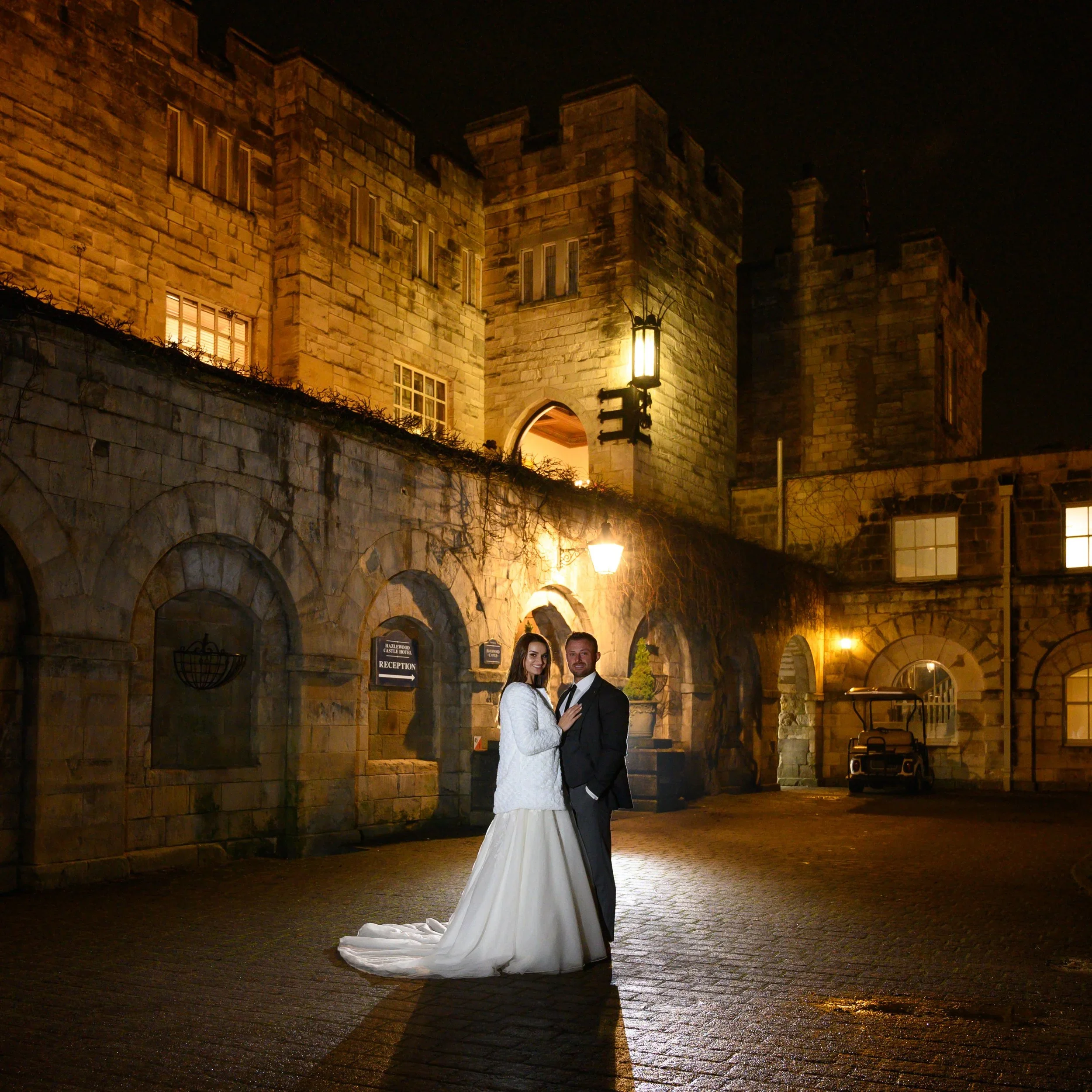 A newlywed couple in wedding attire standing close together at night outside a castle-like building with stone walls and towers, illuminated by warm yellow lights.