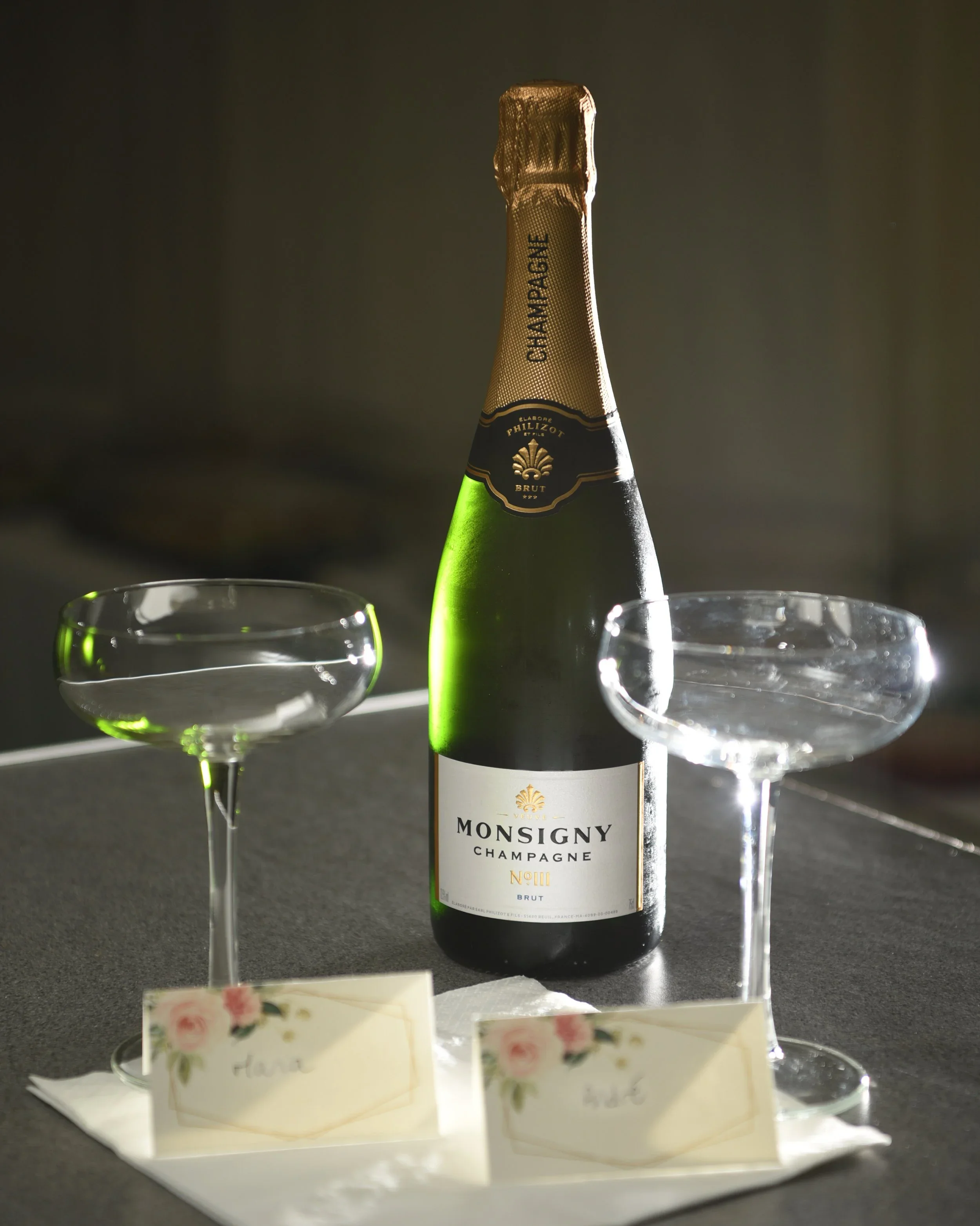 A bottle of Monsigny Champagne Brut with two coupe glasses, one filled with champagne and one empty, on a table with white napkins and name cards.