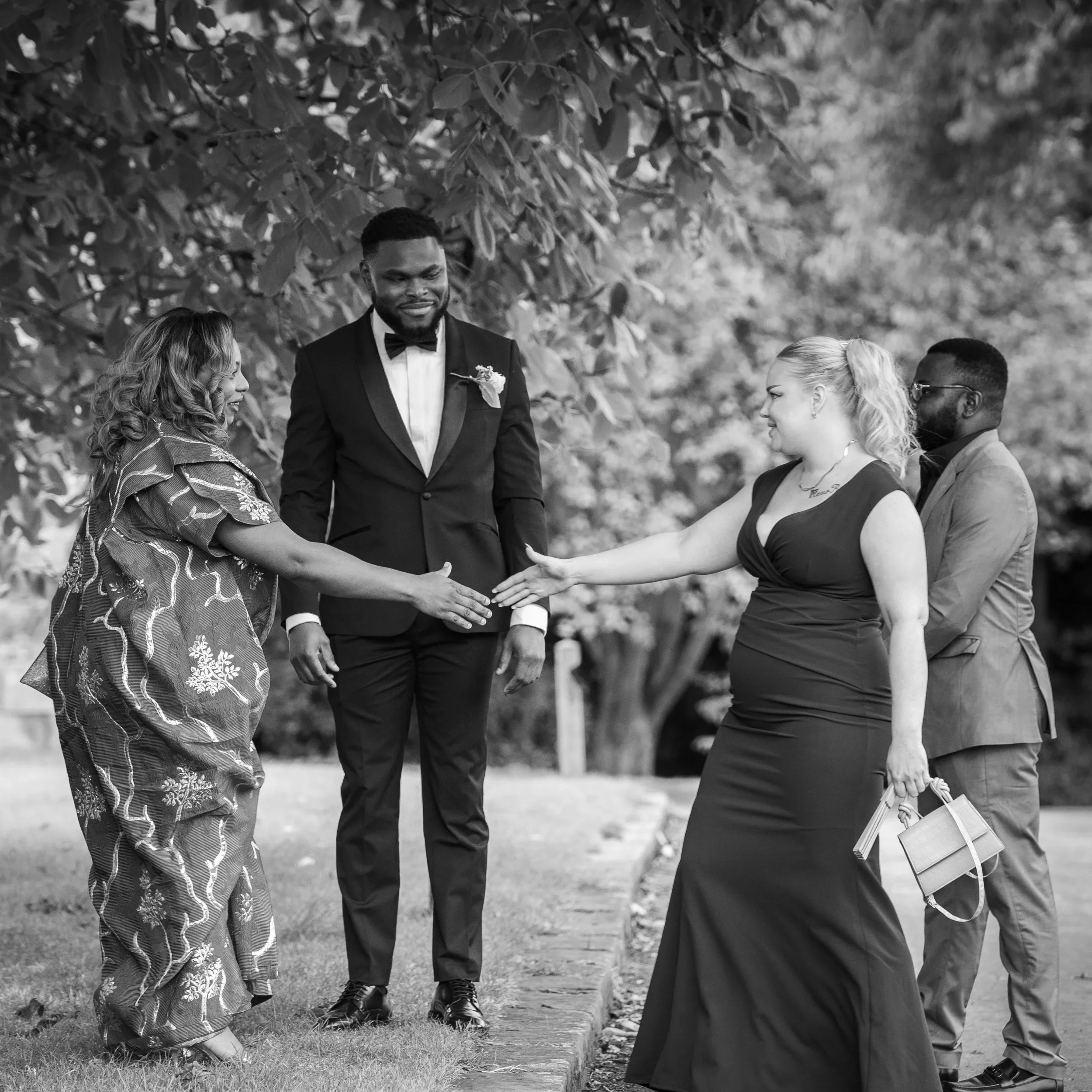 Black and white photo of five people outdoors, two women and three men, greeting each other warmly. One woman in a traditional dress extends her hand to a woman in a black gown, while a man in a tuxedo stands between them. The other man, in a suit and glasses, stands behind the woman in the gown.
