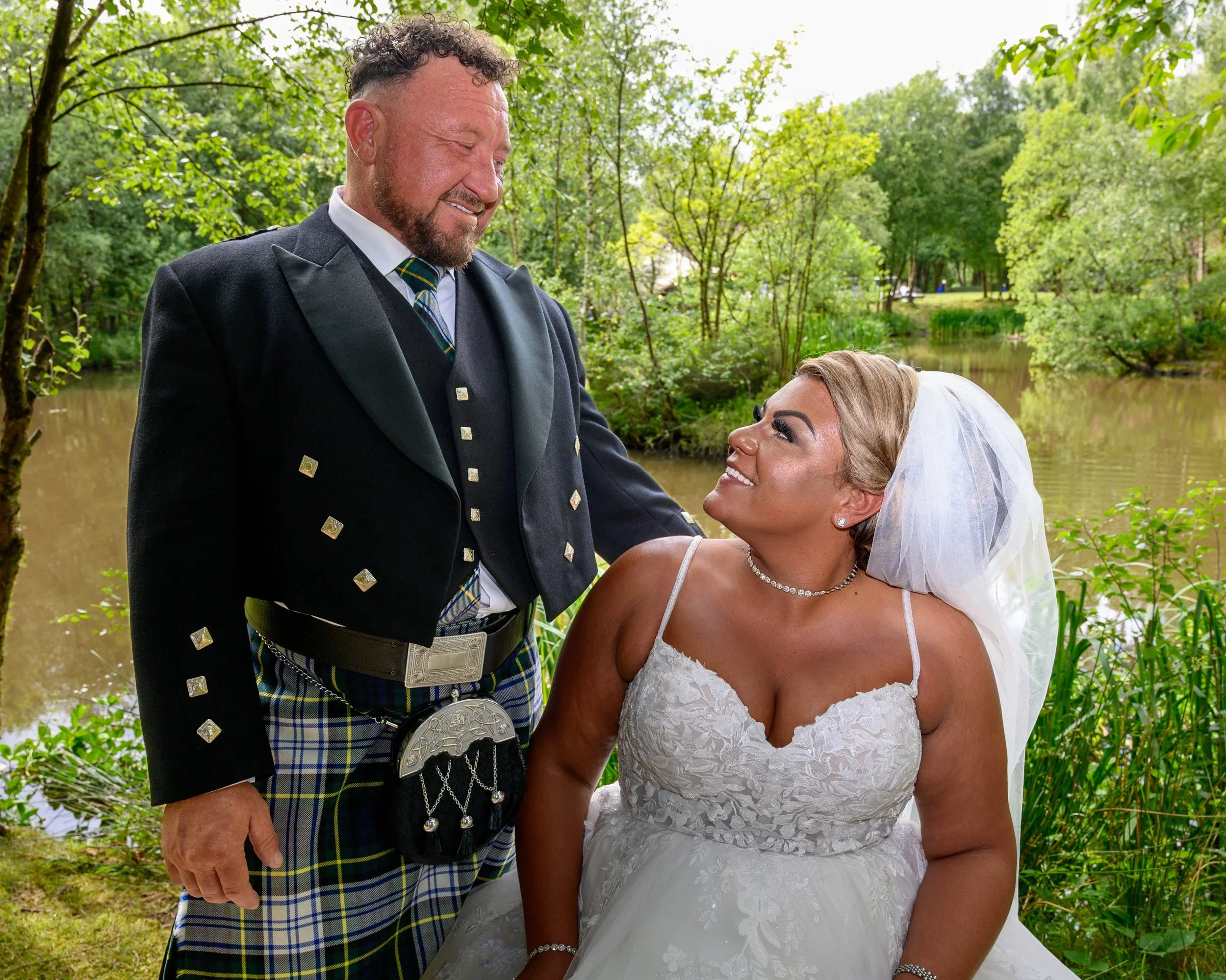 Newlyweds looking at each other. Groom wearing kilt. Seated in front hotel lake at Chevin Country Park Hotel.