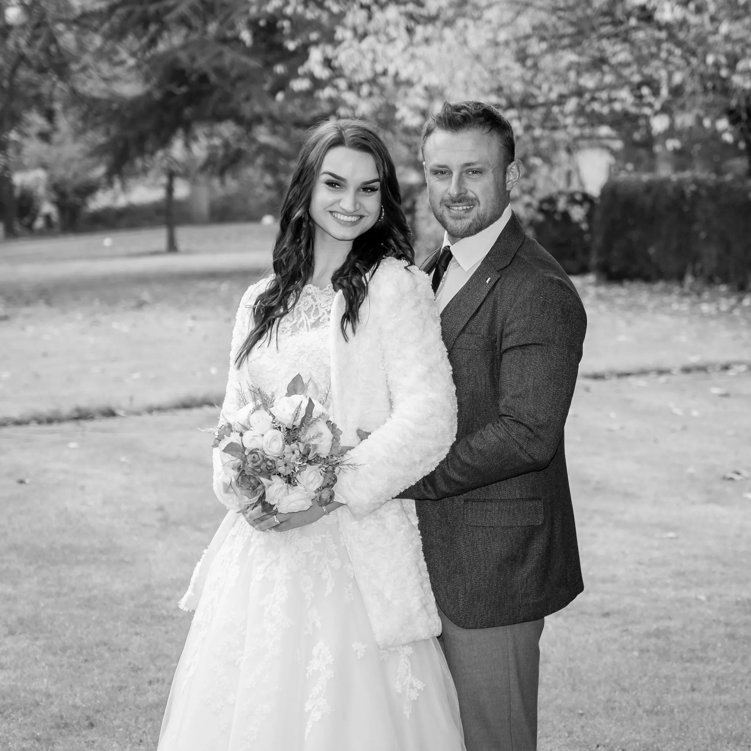 B&W portrait, newlyweds in garden smiling for  camera, Groom stood behind, hands on bride's waist. trees and grass in the background.