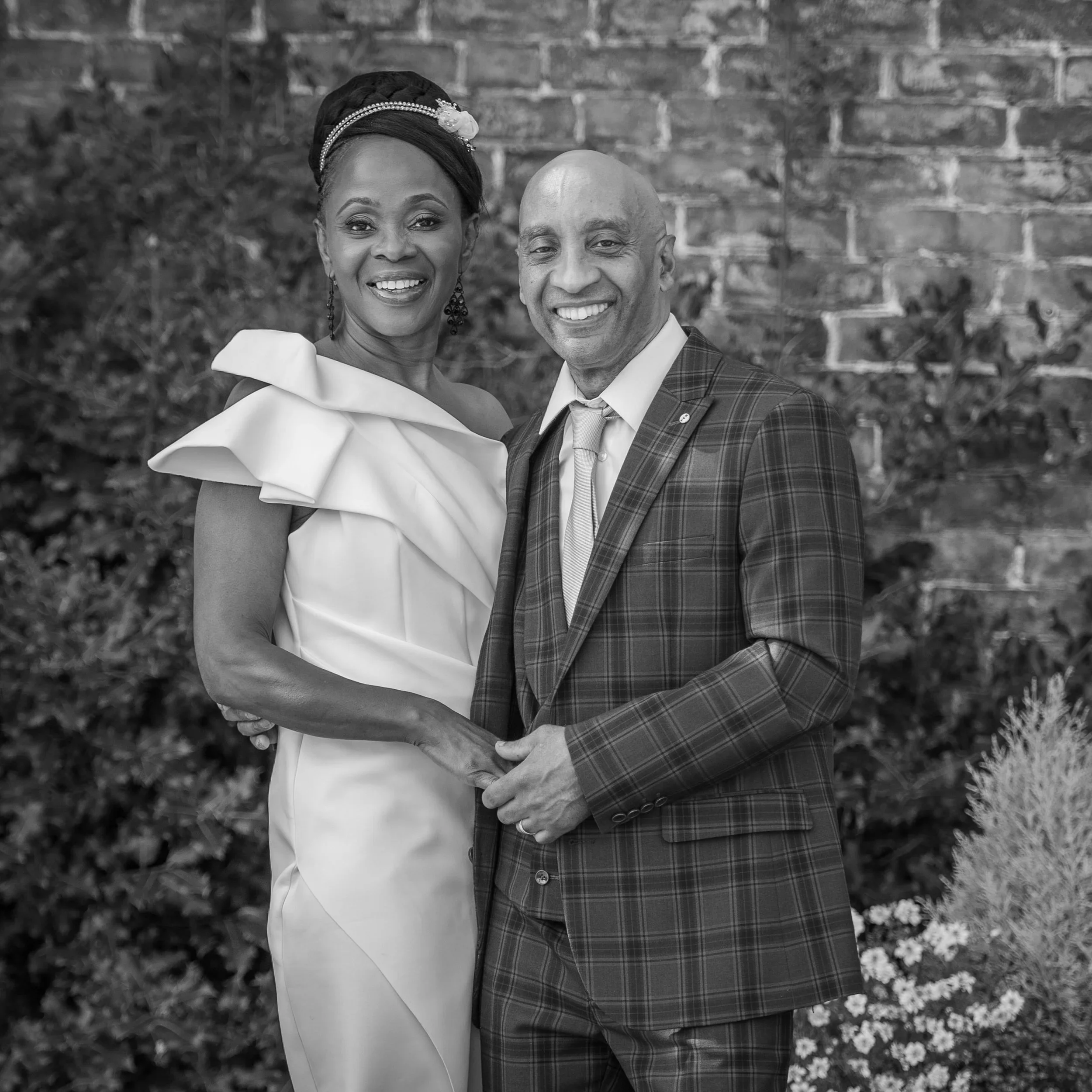 Happy couple smiling, holding hands outdoors, in front of a brick wall. Plants are between them and the wall.