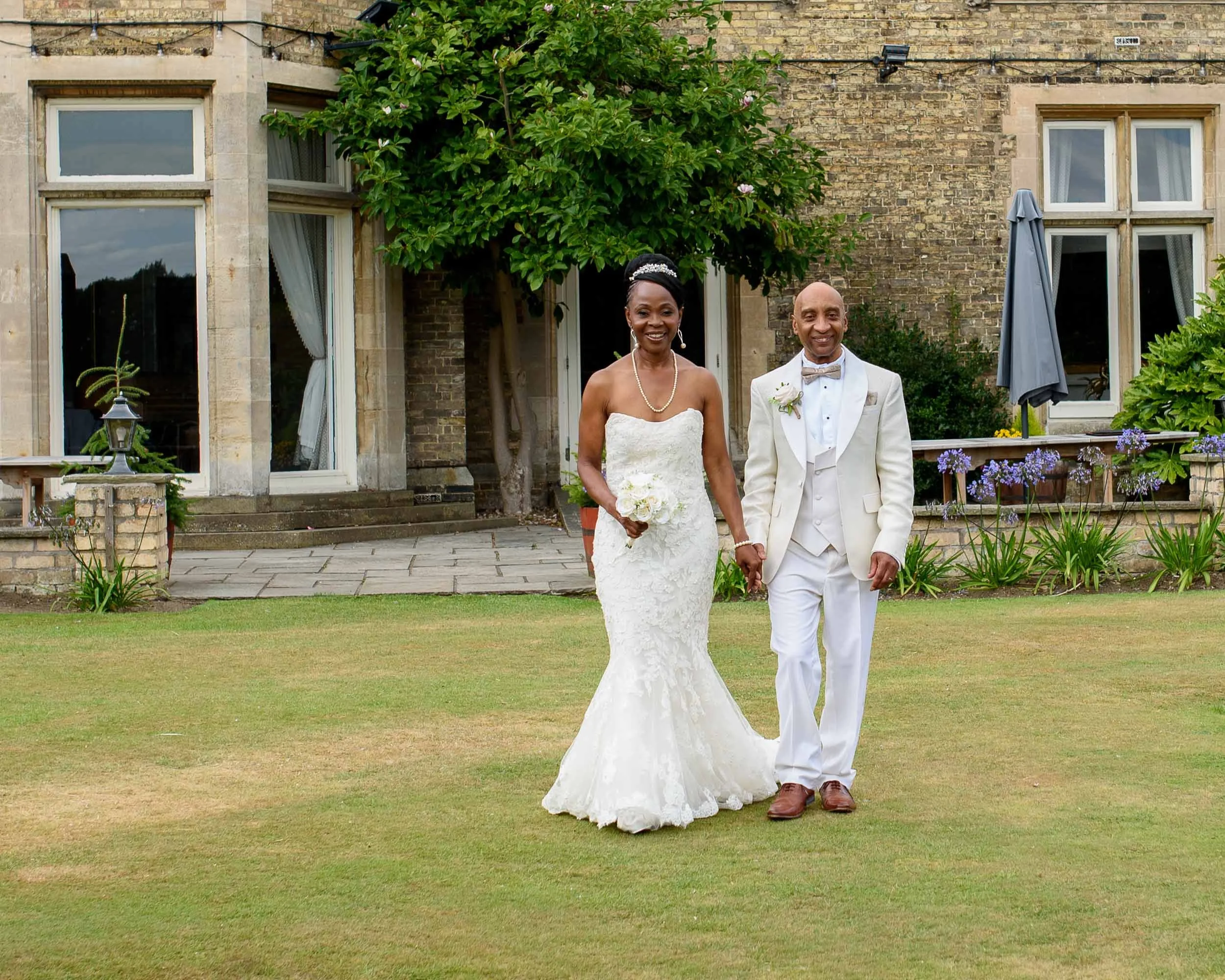 A bride and groom holding hands, smiling, walking on a lawn outside a house. The bride wears a strapless white wedding gown and the groom wears a white tuxedo with a bow tie.