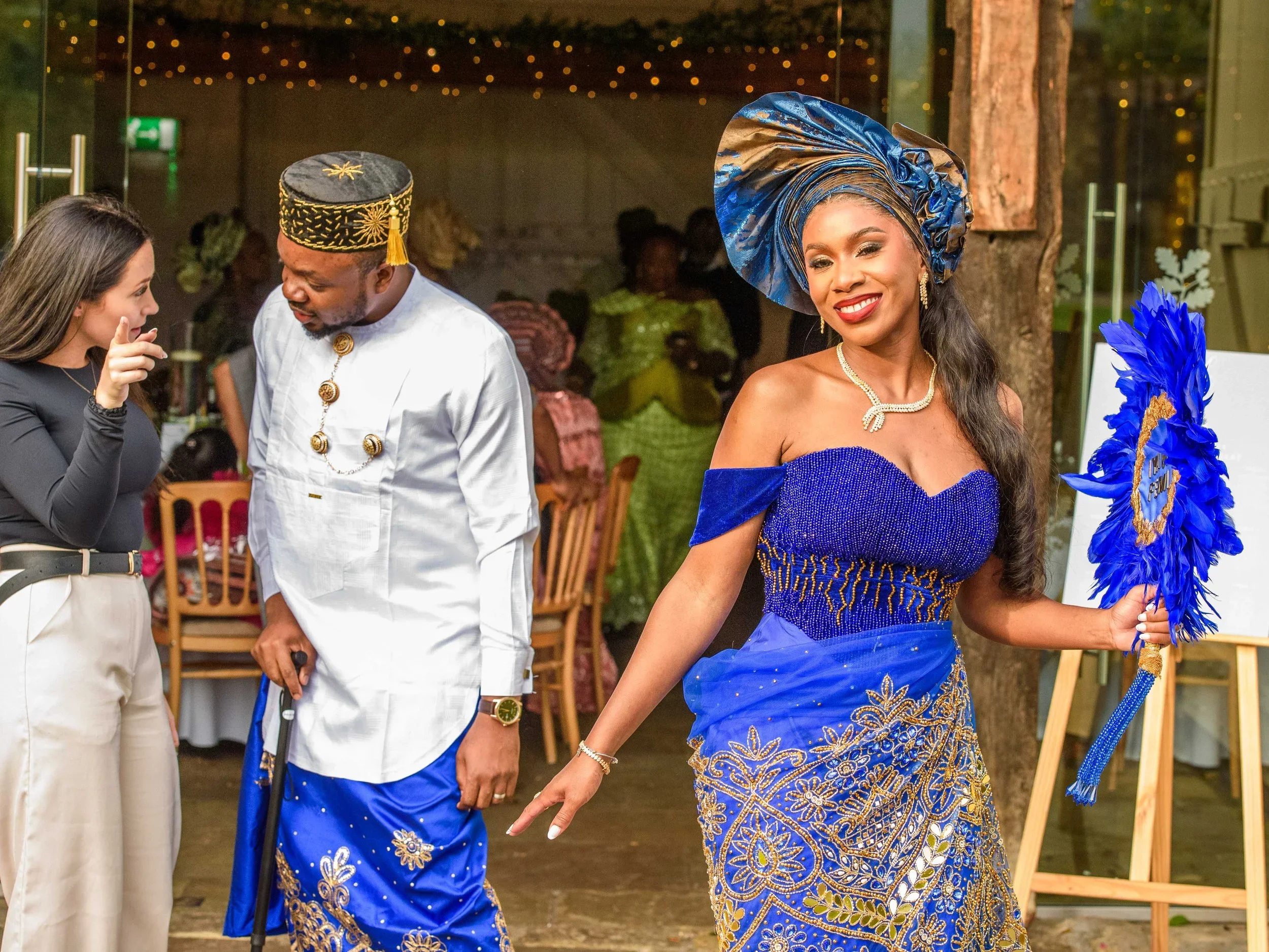 A woman dressed in traditional African attire, wearing a blue and gold dress and a matching head wrap, is smiling and holding a blue feathered fan. She is at a social gathering with other people in the background.