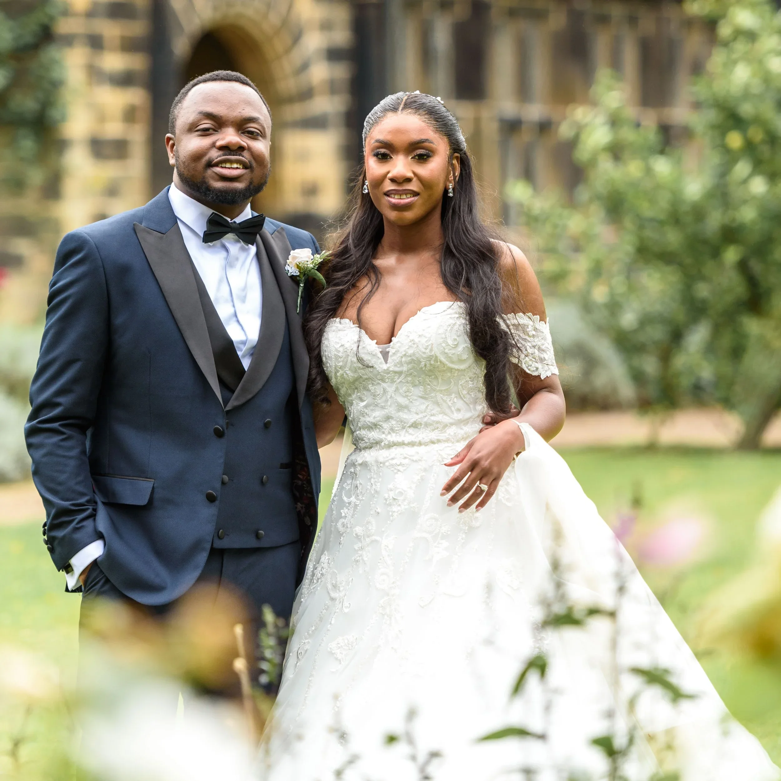 Colour portrait, Bride and groom standing outdoors in front of a historic stone building, dressed in wedding attire, smiling and looking at the camera.