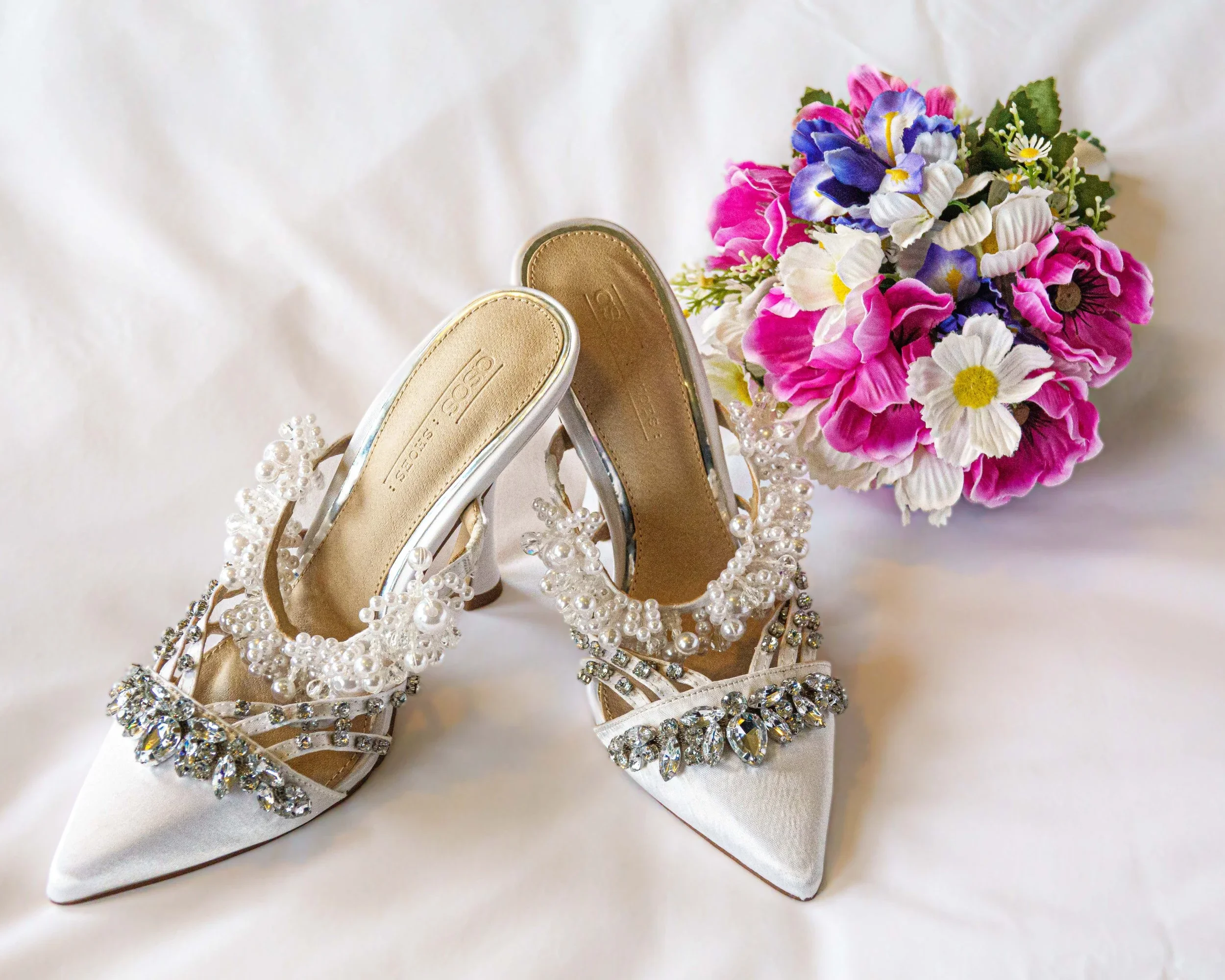 White bridal high heels with pearl and rhinestone embellishments, a bouquet of pink, purple, white, and yellow flowers, all set on a white background.