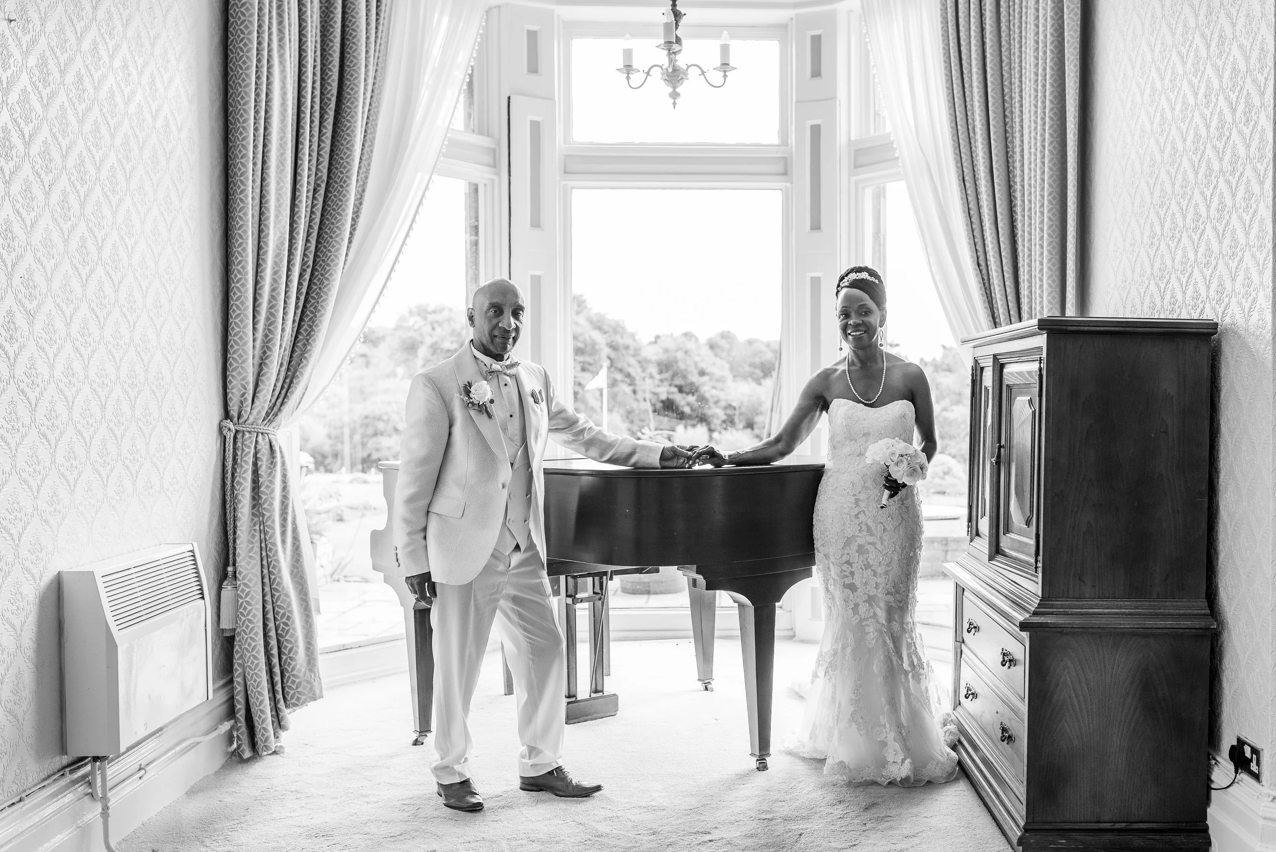 A black and white photo of a bride and groom standing in front of a grand window with curtains, holding hands over a piano, smiling. The groom is dressed in a suit with a boutonniere, and the bride is in a strapless lace wedding dress holding a bouquet.