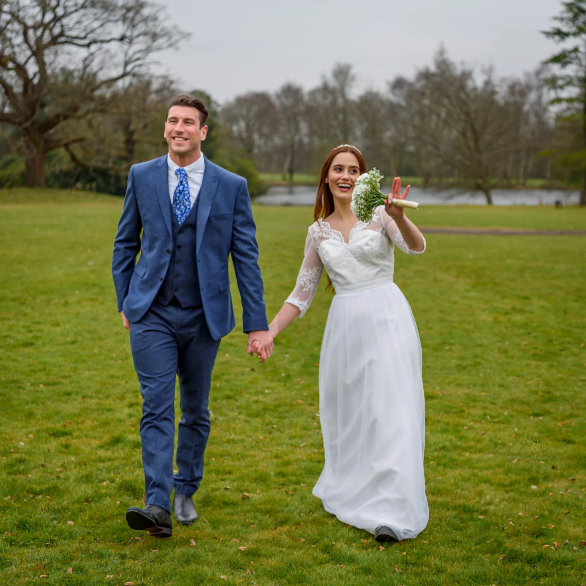 Bride and blue suited groom walking hand in hand through scenic grounds. Bride holding bouquet. Lake in the distance.