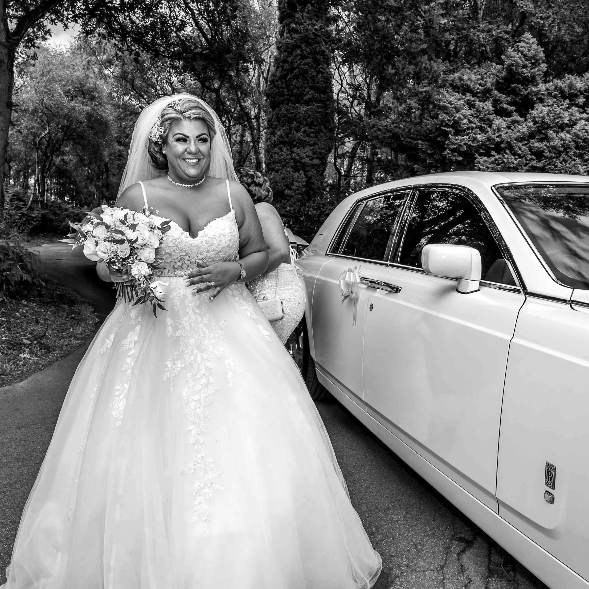 A smiling bride in a wedding gown holding a bouquet of flowers standing next to a luxury car decorated with ribbons on the side mirror, outdoors with trees in the background.