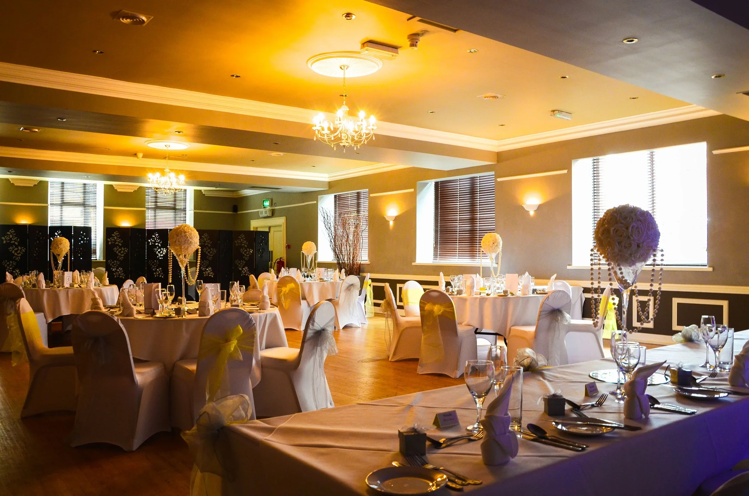 Elegant banquet hall decorated for an event, with round tables covered in white tablecloths, floral centerpieces, and chairs with white covers and bows. Soft lighting and chandeliers hang from the ceiling.