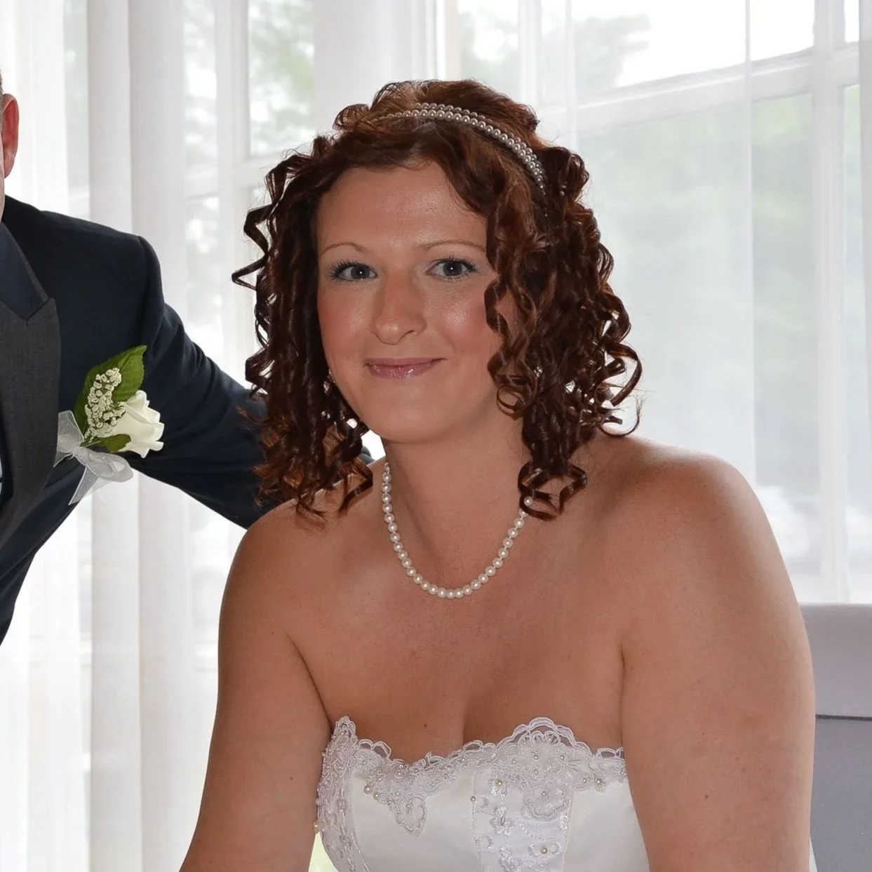A woman in a wedding dress with curly brown hair, wearing a pearl necklace and headband, smiling at the camera, with a man partially visible beside her.