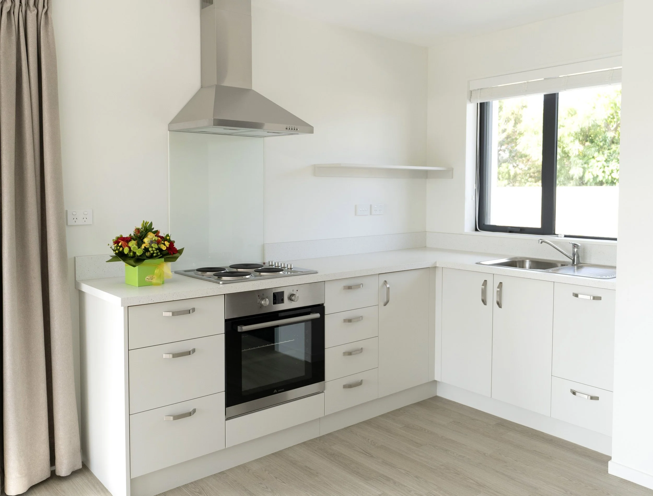 Interior kitchen with bench-tops, oven, rangehood, sink area and exterior window view and drapes