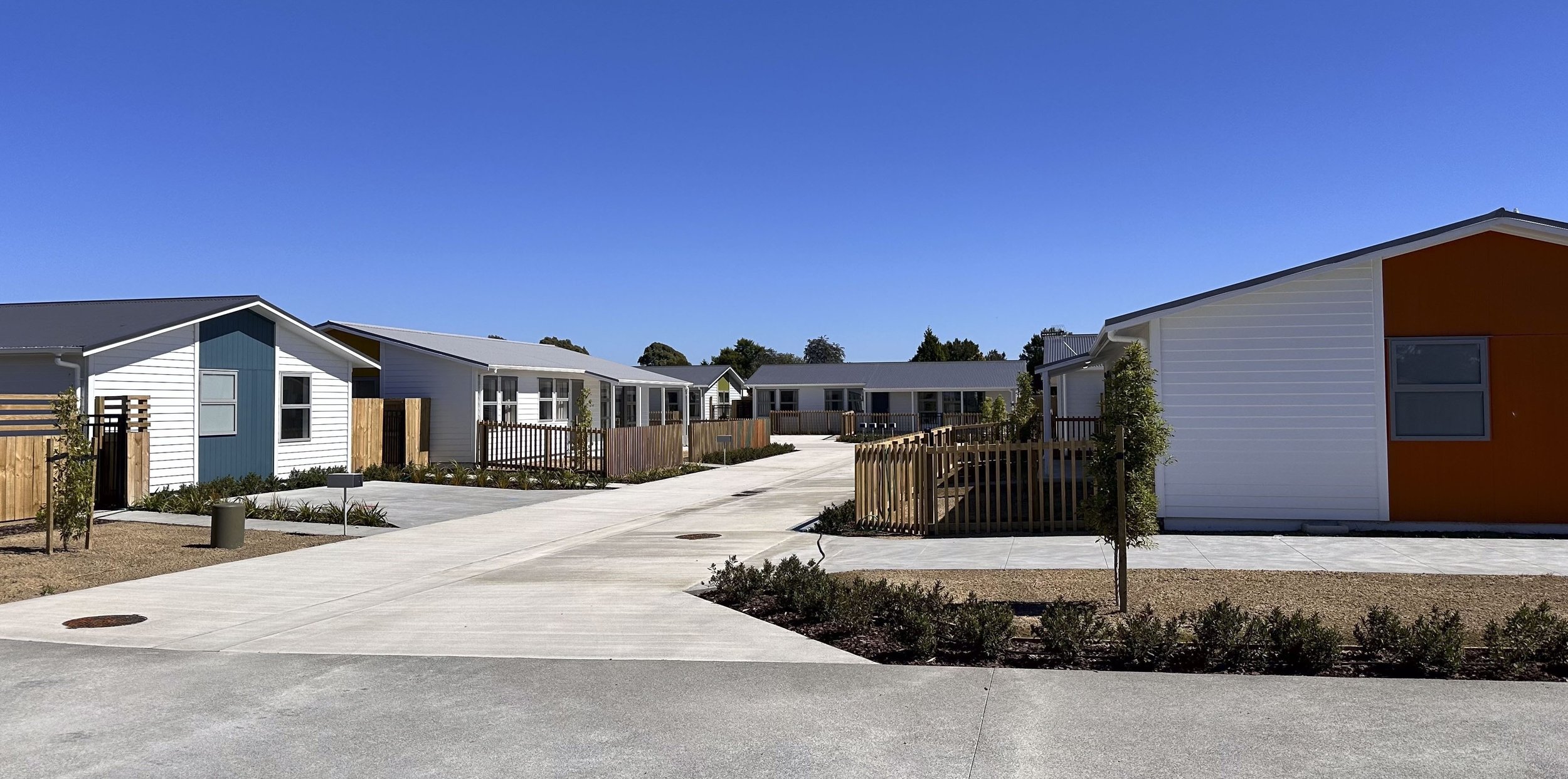 Street-front view up concrete driveway to single-level dwellings showing fencing and planting.