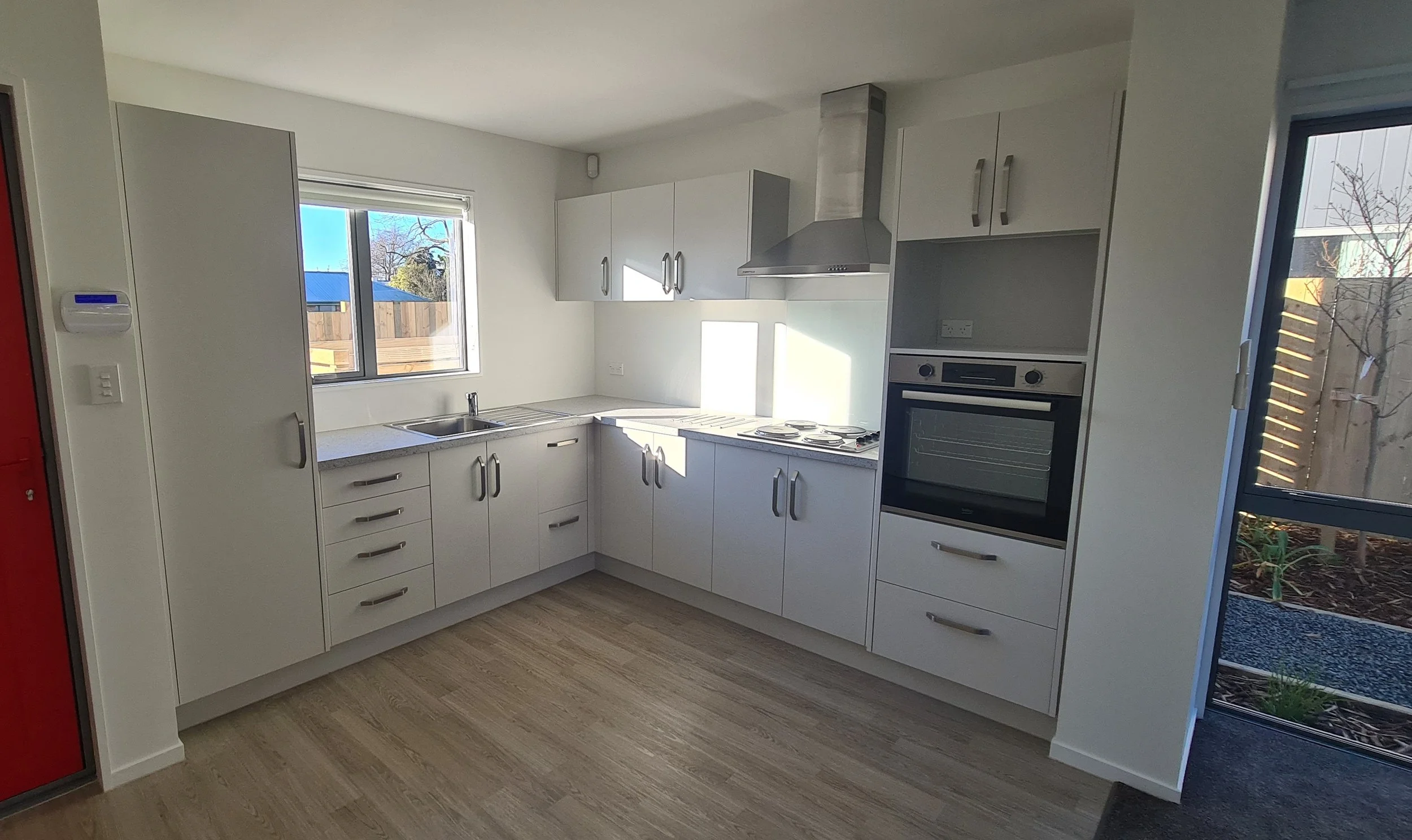 Interior kitchen with bench-tops, oven, rangehood, sink area and exterior window view and doorway
