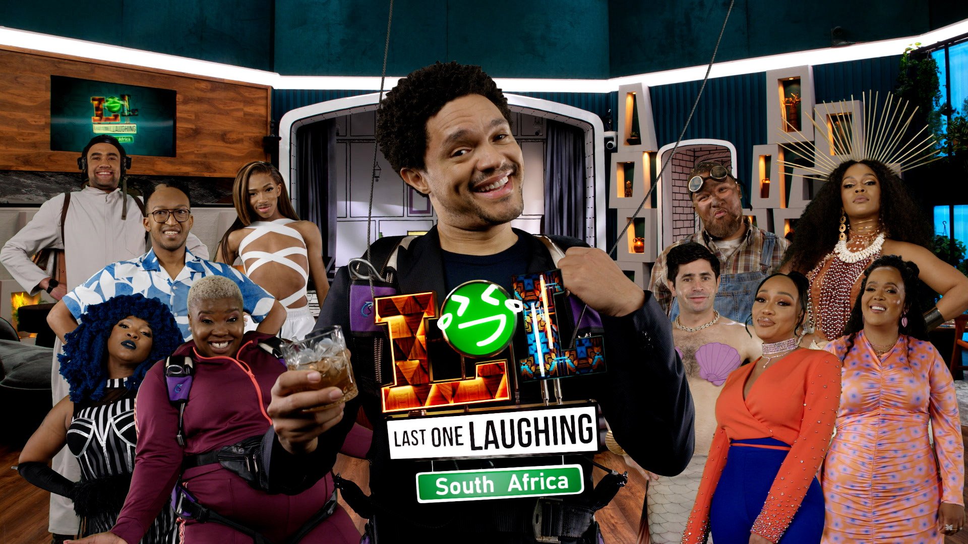 Group of diverse people posing for a photo in a modern room, some holding drinks, with a man in front wearing a sign that reads 'Last One Laughing South Africa.'