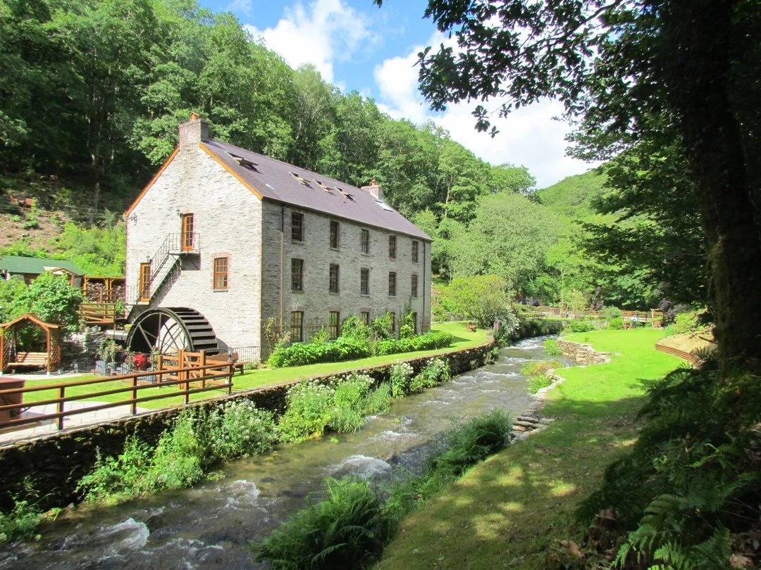 A stone mill house, dog friendly accommodation in Wales  by a flowing river, surrounded by lush green trees and grass on a sunny day.