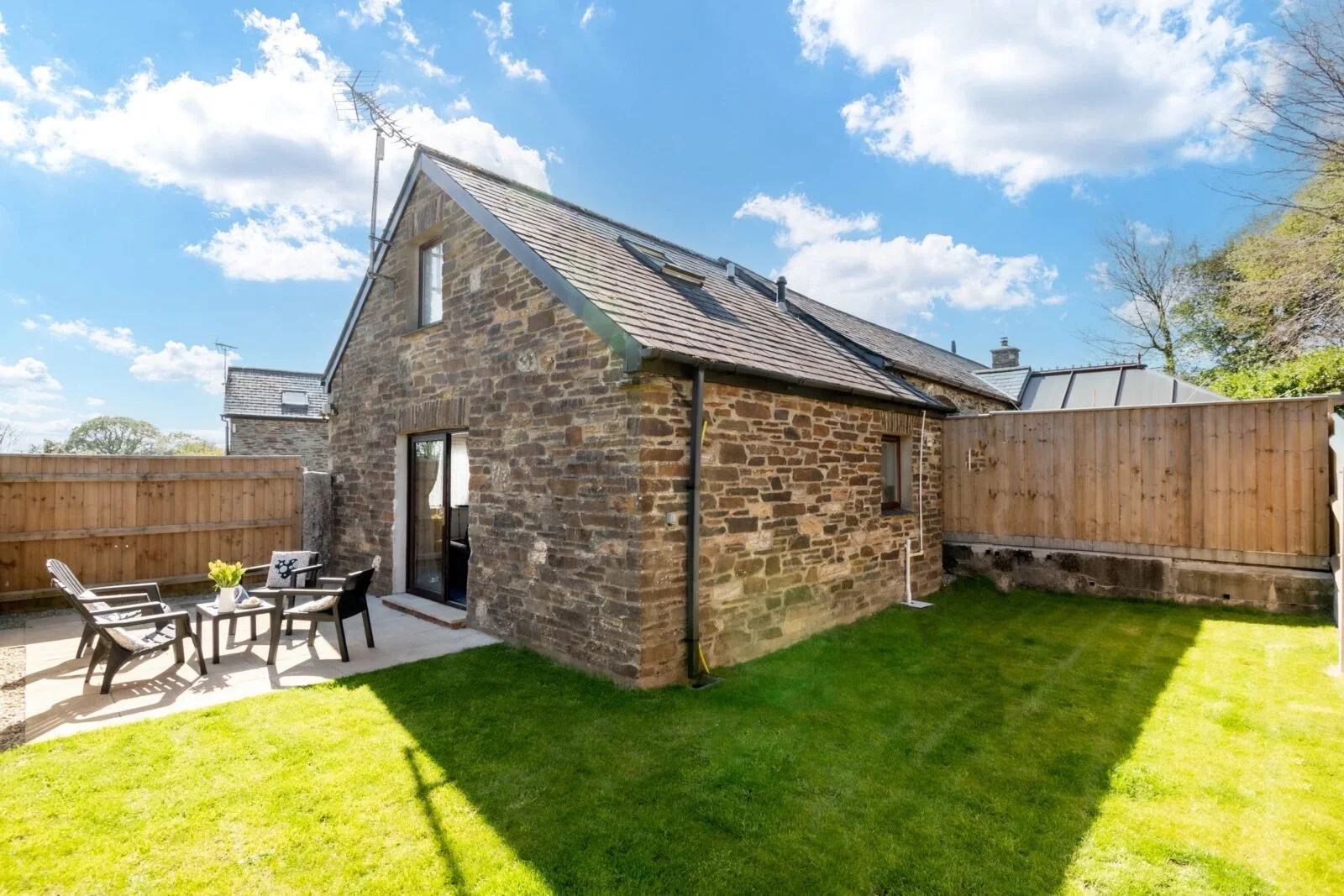 A backyard with a brick house, a small patio area with four black and white chairs and a table with a yellow flower, a well-manicured grass lawn, a wooden fence, and a blue sky with some clouds.