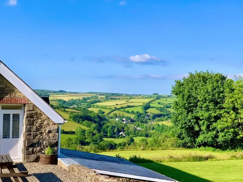 Scenic view of rolling green hills and farmland with a stone house and large tree in the foreground on a sunny day.